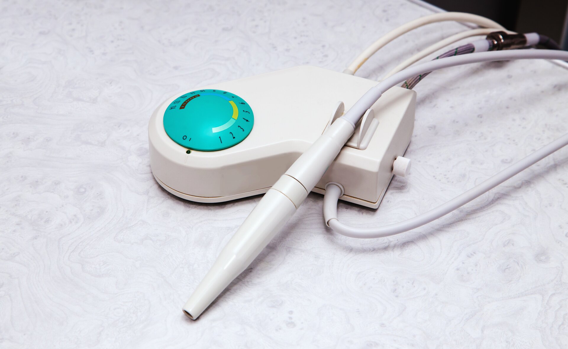 Na zdjęciu zaprezentowano skaler ultradźwiękowy. Urządzenie składa się z modułu sterującego, który mieści się w niewielkiej prostokątnej skrzynce, do której został podłączony przewód zakończony rączką. Na module znajduje się również pojemnik na wodę, a także pokrętła i przyciski sterujące pracą skalera.