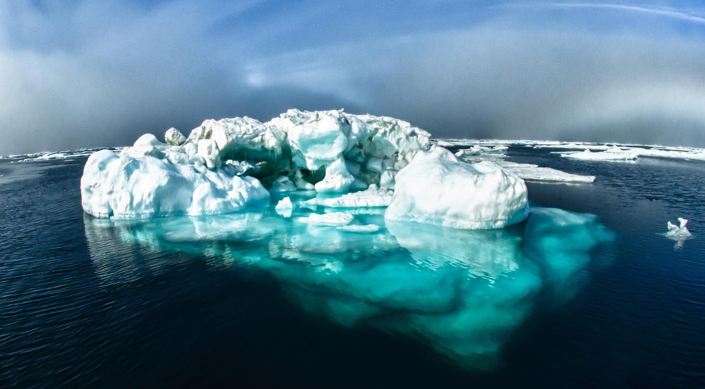 Fotografia przedstawia fragment oceanu . W części środkowej zdjęcia znajduje się góra lodowcowa w połowie zanurzona pod wodą.