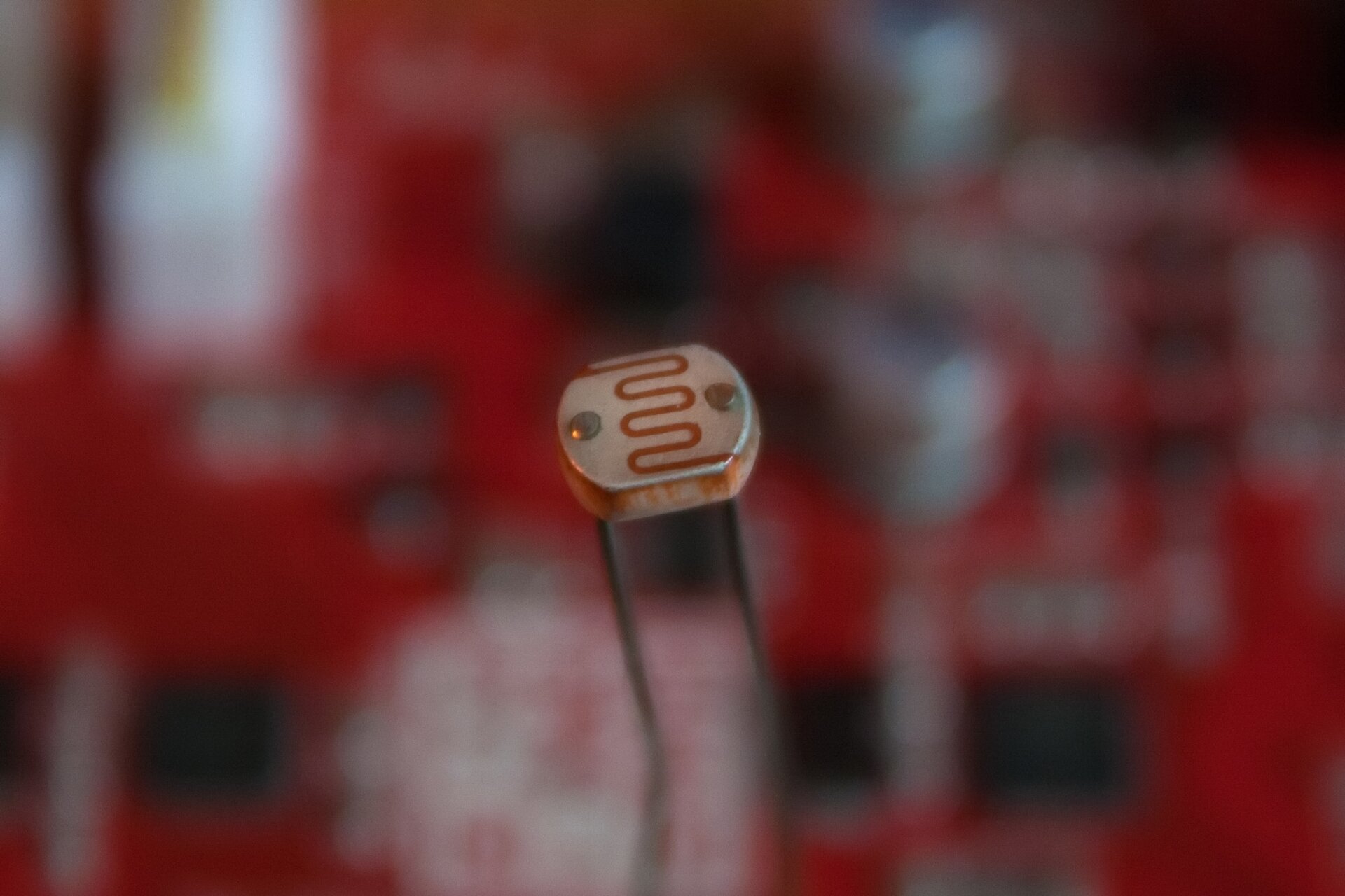 Rys. 1. przedstawia fotografię fotorezystora. Na rozmytym tle w barwach czerwono‑szarych widnieje metalowy przedmiot w kształcie małej srebrnej blaszki z narysowaną krzywą przypominającą sinusoidę. Z dolnej części blaszki odchodzą 2 pręty metalowe w kolorze ciemnoszarym.