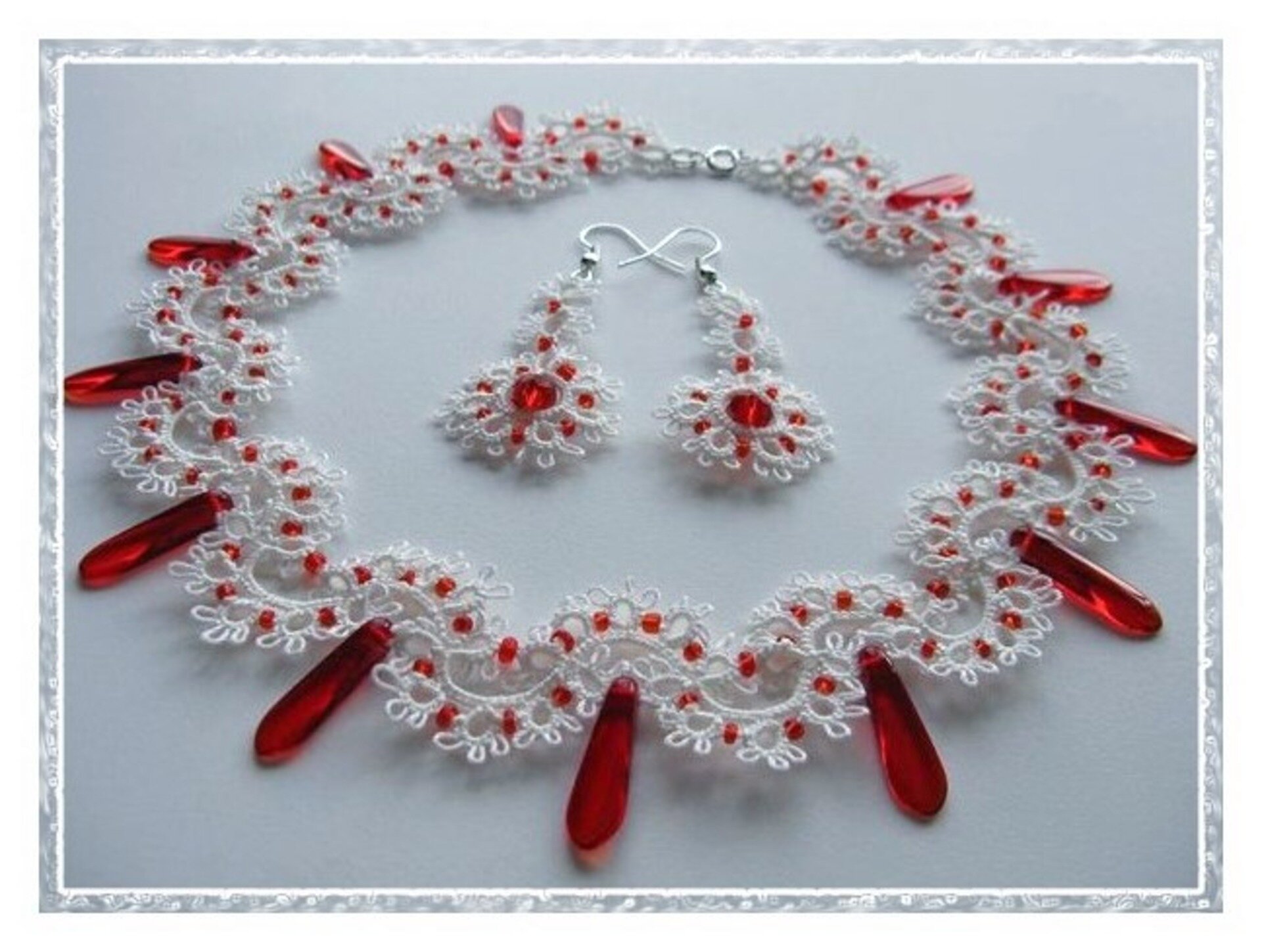 Fotografia przedstawia wyrób rękodzielniczy – naszyjnik i kolczyki. Zestaw zrobiono z połączenia białej koronki frywolitkowej i czerwonych koralików.