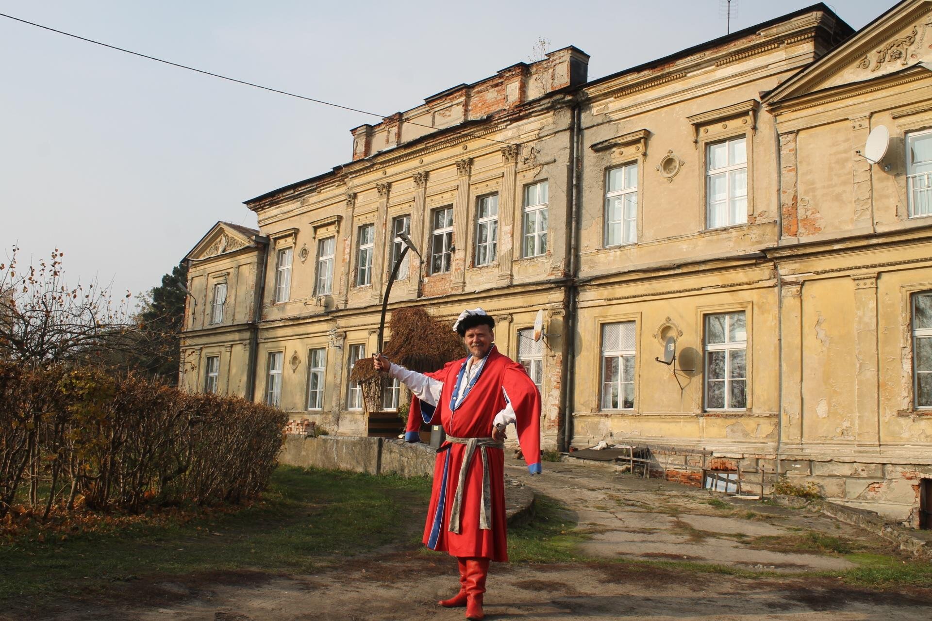 Zdjęcie przedstawia żółty, poniszczony budynek, na tle którego stoi mężczyzna w czerwonej szacie. Budynek jest piętrowy, symetryczny, a umieszczone na nim okna są ułożone w równych odległościach. Przed dworkiem widoczne są krzewy z pomarańczowymi liśćmi oraz zieloną trawę porastającą przestrzeń pomiędzy. Mężczyzna znajdujący się na pierwszym planie ubrany jest w czerwoną, sięgającą mu do kostek, szatę oraz czerwone, skórzane kozaki. W pasie przewiązany ma żółty pas z niebieskimi elementami. Pod szatą widoczna jest biała koszula. Na głowie ma czarno‑białą czapkę. Mężczyzna trzyma lewą rękę na biodrze, a prawą unosi ku górze. Trzyma w niej czarny kij, zakończony srebrnym narzędziem. Niebo jest błękitne, bezchmurne. Zdjęcie zostało wykonane w świetle dziennym.