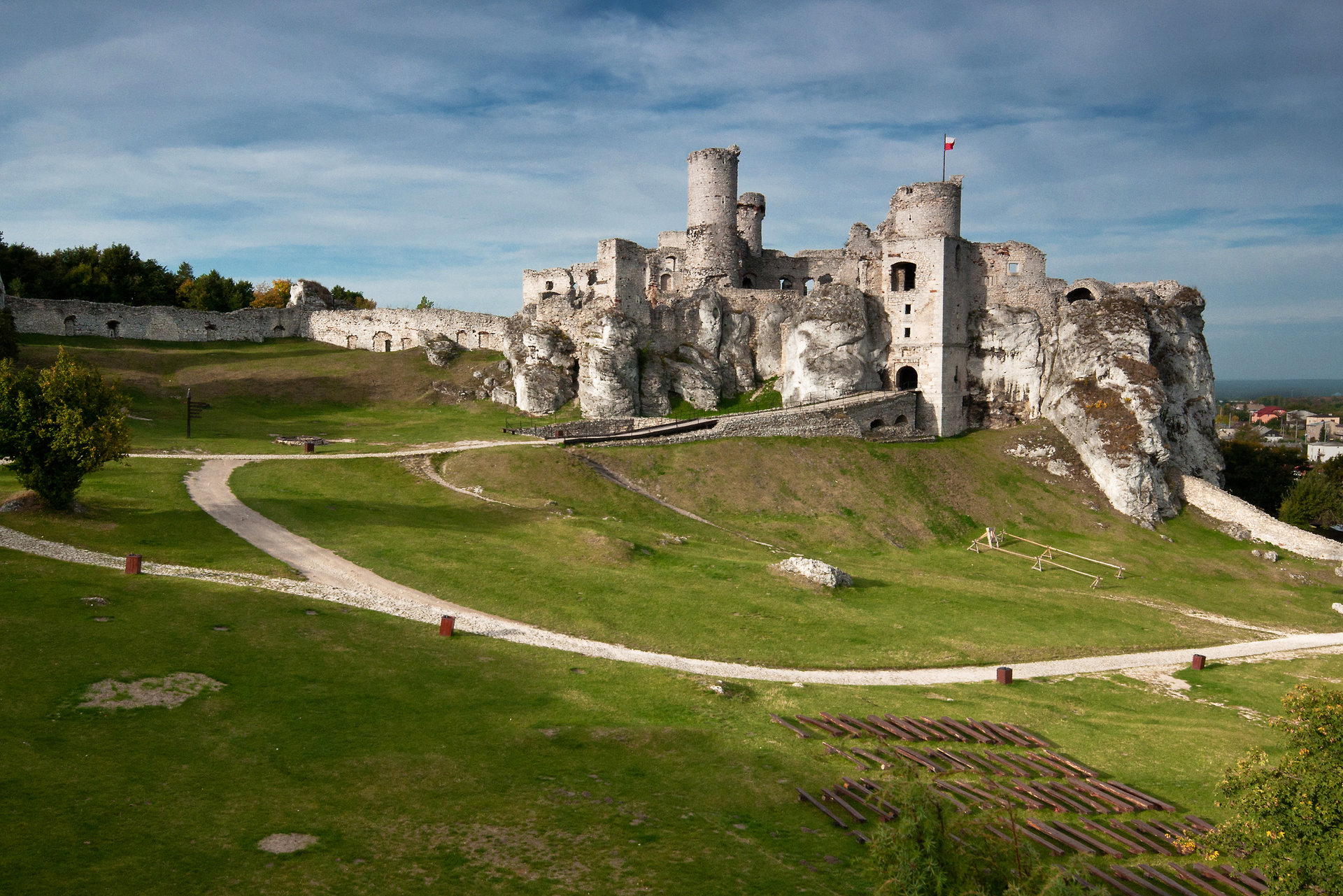 Fotografia prezentuje ruiny zamku Ogrodzieniec, umiejscowione na wzgórzu, do którego prowadzi jasna, kręta droga. Fragment zamku z prawej strony zdjęcia wbudowany jest w naturalne skały. Zamek zbudowany jest z jasnego kamienia - z piaskowca.