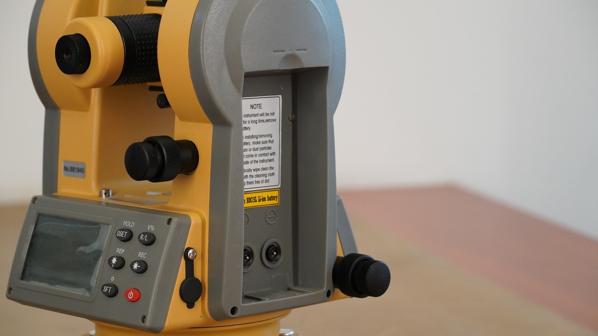 Zdjęcie przedstawia widok teodolitu z bocznej perspektywy. Widać na nim miejsce, w którym umieszcza się baterie. Zdjęto z niego osłonę. Po lewej stronie widoczny jest okular, a pod nim wyświetlacz z przyciskami.