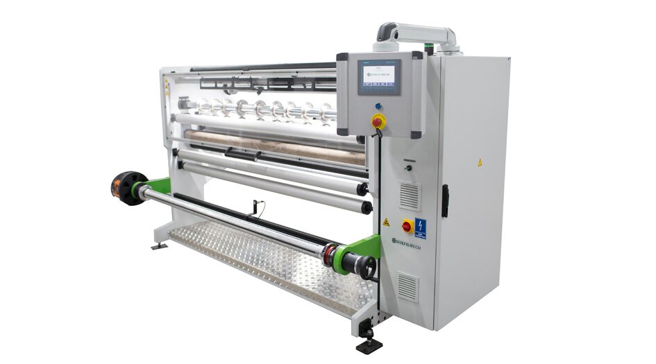 Zdjęcie ukazuje bobiniarkę. Urządzenie ma kształt dużego prostopadłościanu. W przedniej części widoczny jest wałek do nawijania papieru, a wyżej cały system wałków i ostrzy. Na prawo znajduje się moduł zasilający z panelem do sterowania urządzenia.