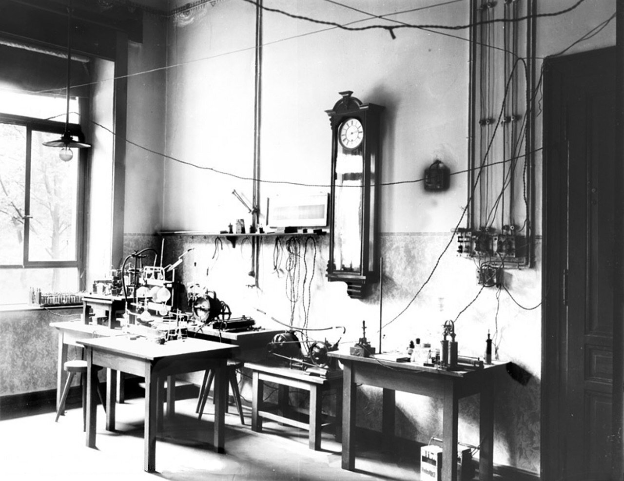 Zdjęcie przedstawia pracownię, a ściślej mówiąc laboratorium Wilhelma Roentgena. Przestrzenne wnętrze z wysokim sufitem, pod ścianą stoły, lampy, kable, szklane naczynia. 