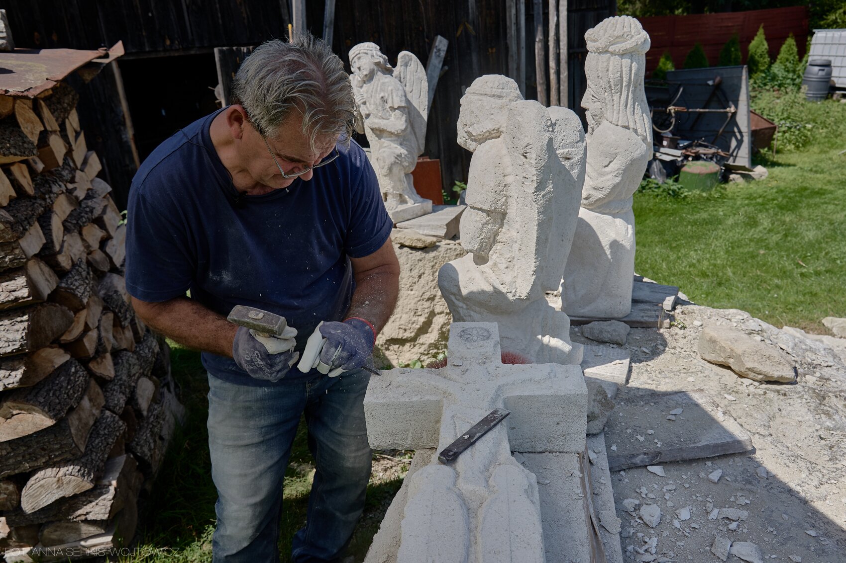 Ilustracja interaktywna przedstawiająca rzeźbiarza w pracy. Jest to siwowłosy mężczyzna w okularach, ubrany w jeansy oraz granatową koszulkę. Ma na sobie rękawice, jego wzrok skierowany jest w dół, na kamień oraz narzędzia, które trzyma w dłoniach. Tło stanowi sterta drewna uchwycona z lewej strony, fragment drewnianego budynku gospodarskiego oraz trawa.