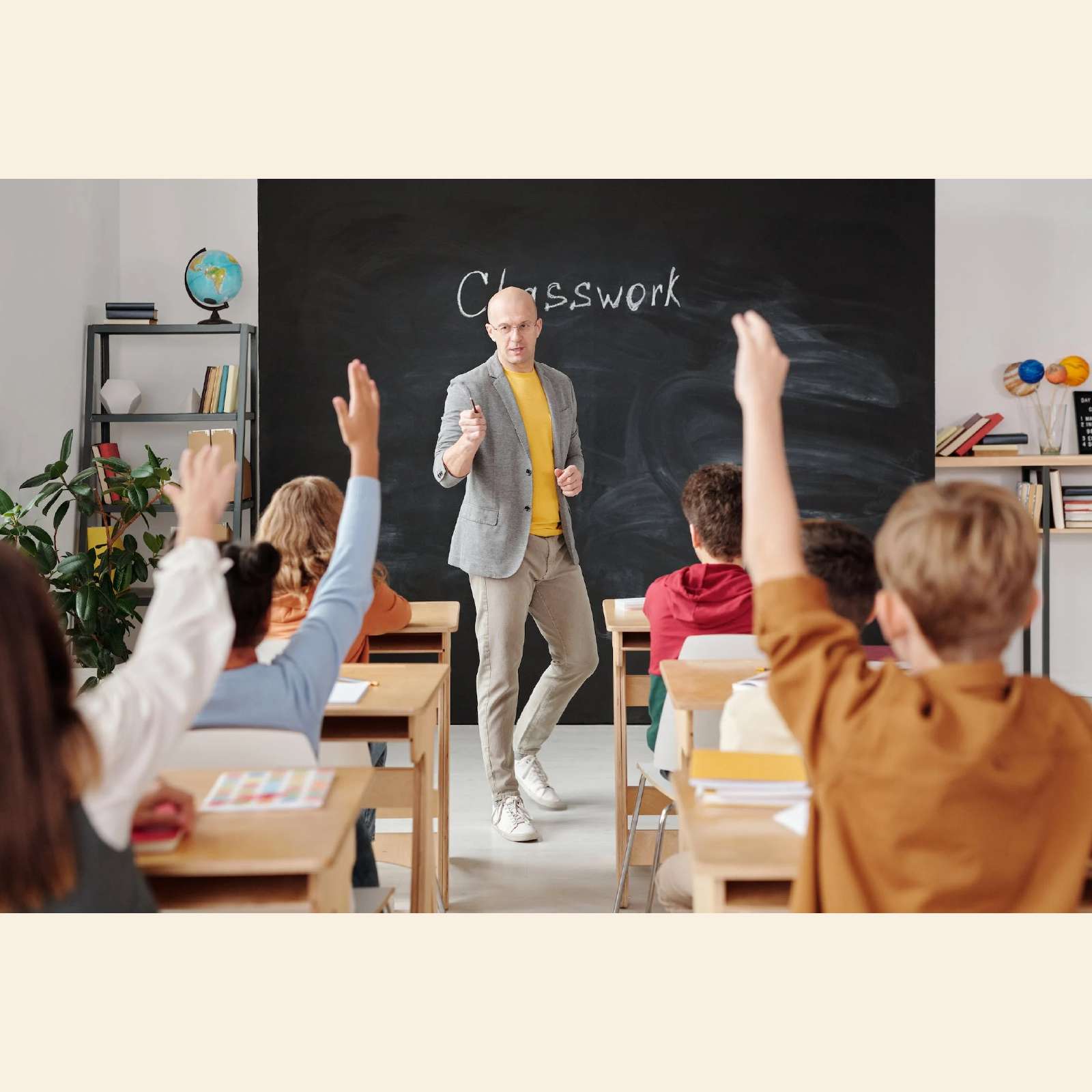 Zdjęcie przedstawia salę klasową w szkole. Widoczni są uczniowie w ławkach oraz nauczyciel stojący przed tablicą. Uczniowie podnoszą ręce do odpowiedzi. 