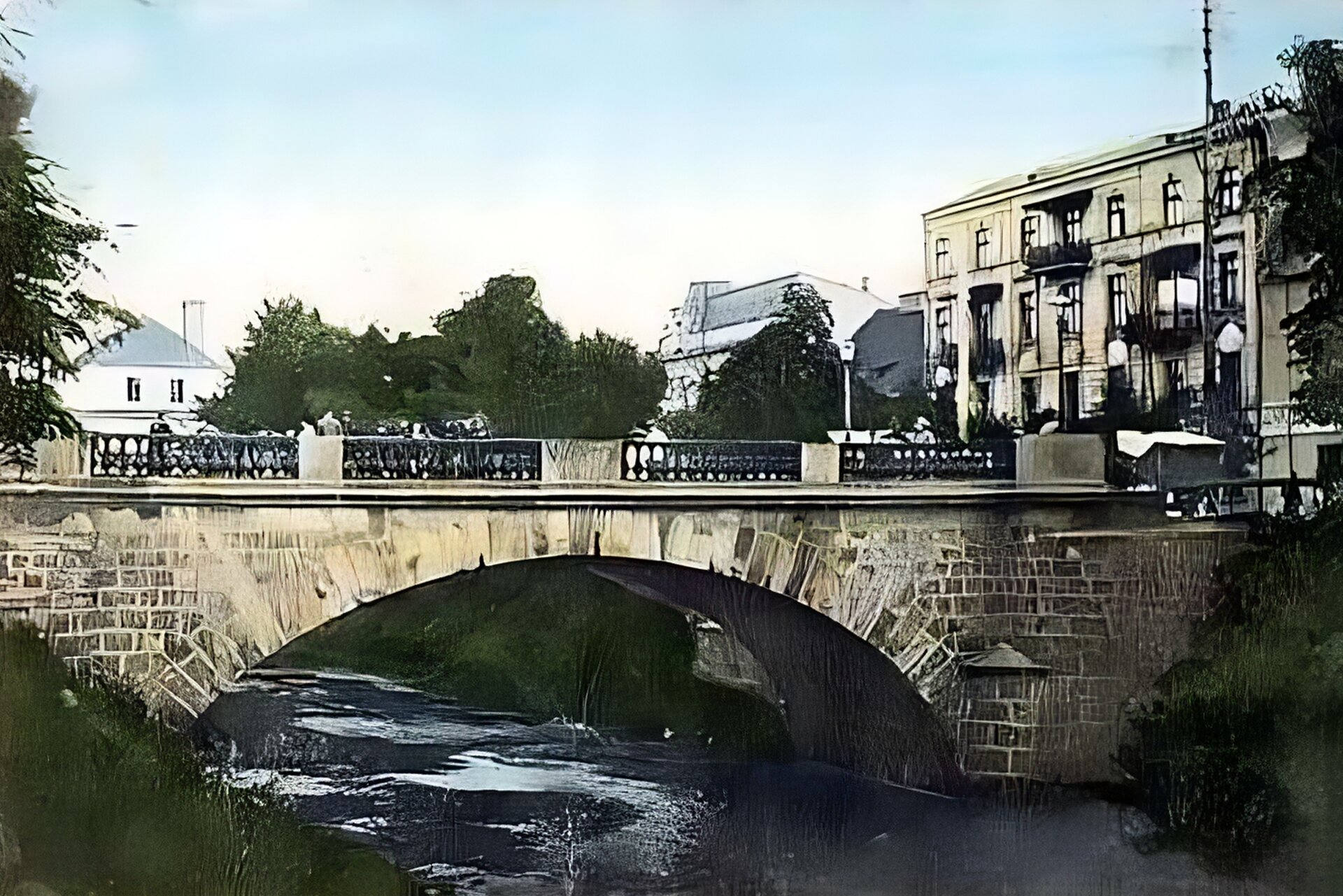 Zdjęcie przedstawia nieduży, kamienny most łukowy nad rzeką. Most zbudowany z szarych, prostokątnych kamieni. Przez całą długość mostu przechodzi balustrada z pękatymi tralkami. W tle widać budynki. Na brzegach rzeki rośnie zielona trawa.