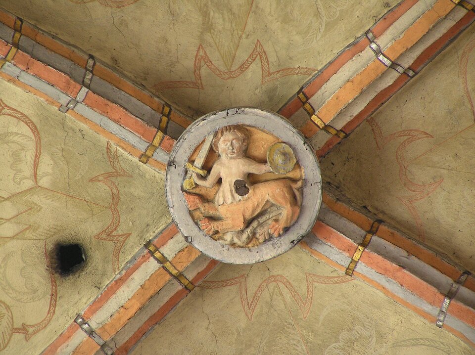 Ilustracja przedstawia zwornik, na którym umieszczony jest wizerunek hybrydy człowieka i zwierzęcia. Prawdopodobnie jest to postać fauna. W jednej z dłoni trzyma miecz, a w drugiej tarczę.