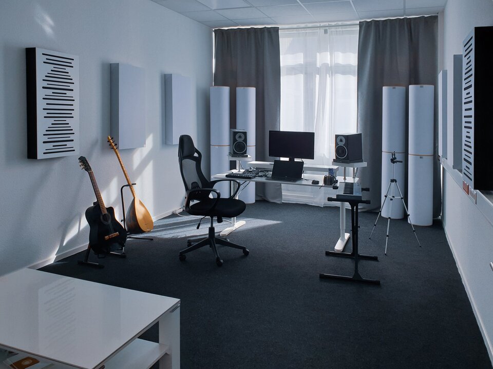 Niewielkie studio do produkcji muzyki Pomieszczenie na zdjęciu ma jasne ściany i sufit oraz grafitową podłogę. W lewym dolnym rogu znajduje się biały niski stolik z półką pod blatem. Na środku jest czarne krzesło na kółkach, keyboard na czarnym stojaku oraz białe biurko. Na biurku leży czarny laptop, za nim na podwyższeniu stoi czarny monitor. Po bokach monitora, również na podwyższeniu, stoją kolumny głośnikowe w kształcie prostopadłościanu. Mają po dwa okrągłe głośniki. Dolny głośnik jest czarny z jasną ramką, a górny jasny z małym, czarnym trójkątem w środku. Po lewej stronie laptopa leżą dwie małe konsole muzyczne i kable. Po prawej stronie jest myszka na kablu, czarne słuchawki oraz niebieska puszka. Za krzesłem stoją oparte o czarne stojaki dwa instrumenty. Pierwszy to czarno‑brązowa gitara, a drugi to mandolina w kolorze jasnego drewna. Po prawej stronie biurka stoi srebrno‑czarny trójnogi statyw. W tle znajduje się duże okno z białą firanką i szarymi zasłonami po bokach. Przed zasłonami stoją po dwie duże, białe kolumny. Na ścianach są powieszone po trzy panele w kształcie prostopadłościanu. Jeden biały z czarnymi bokami i czarnymi, poziomymi liniami o różnej długości oraz dwa całe białe.