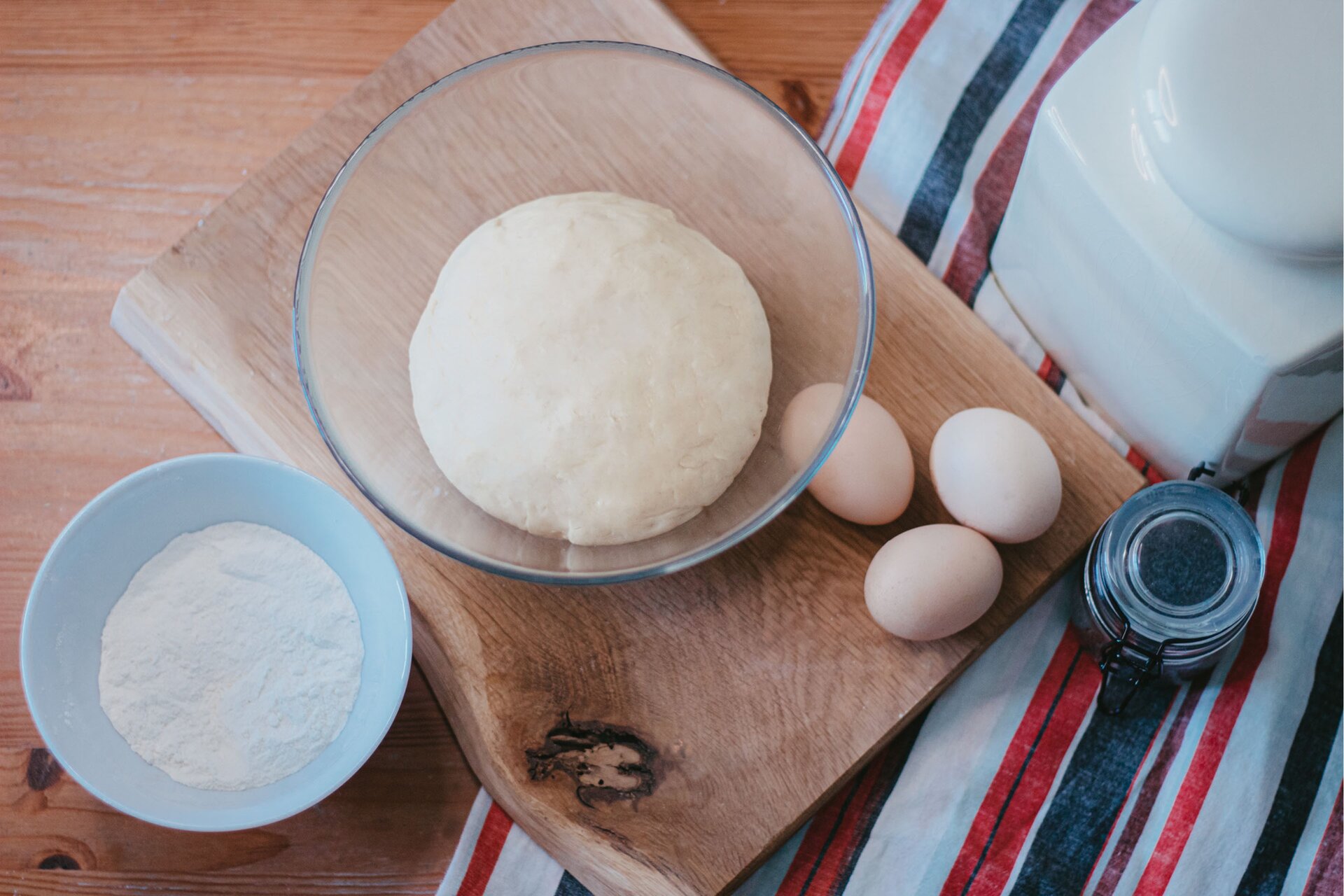 Zdjęcie przedstawia krok pierwszy formowania wianka debrzeńskiego. Ujęcie wykonane jest z góry pod skosem. Widoczny jest jasnobrązowy drewniany blat na którym w środkowej części grafiki znajduje się jasnobrązowa drewniana deska ułożona pod skosem. Na środku deski dostrzec można szklaną przezroczystą dużą miskę w środku której znajduje się jasna kula ciasta. Obok miski w dolnej części grafiki znajdują się trzy jajka. W lewym rogu zdjęcia widoczna jest stojąca na blacie mała biała miska wypełniona do połowy białą mąka. W prawej części zdjęcia znajduje się biała ścierka w czarne i czerwone pasy ułożona pionowo. Na fragmencie deski stoi deska. Na ścierce znajduje się mały szklany przezroczysty słoik z czarnym klipsem wypełniony w środku niebieskim makiem. W prawym górnym rogu zdjęcia widoczny jest biały, wysoki, prostokątny pojemnik.