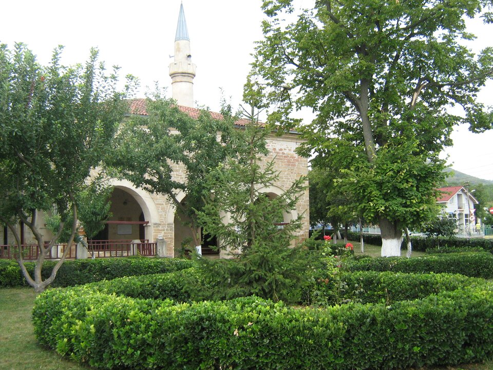 Zdjęcie przedstawia meczet. Jest to murowany budynek z arkadami, pokrytym dachówką dachem oraz minaretem ze strzelistym dachem i głośnikami. Przed budynkiem są drzewa oraz żywopłot.