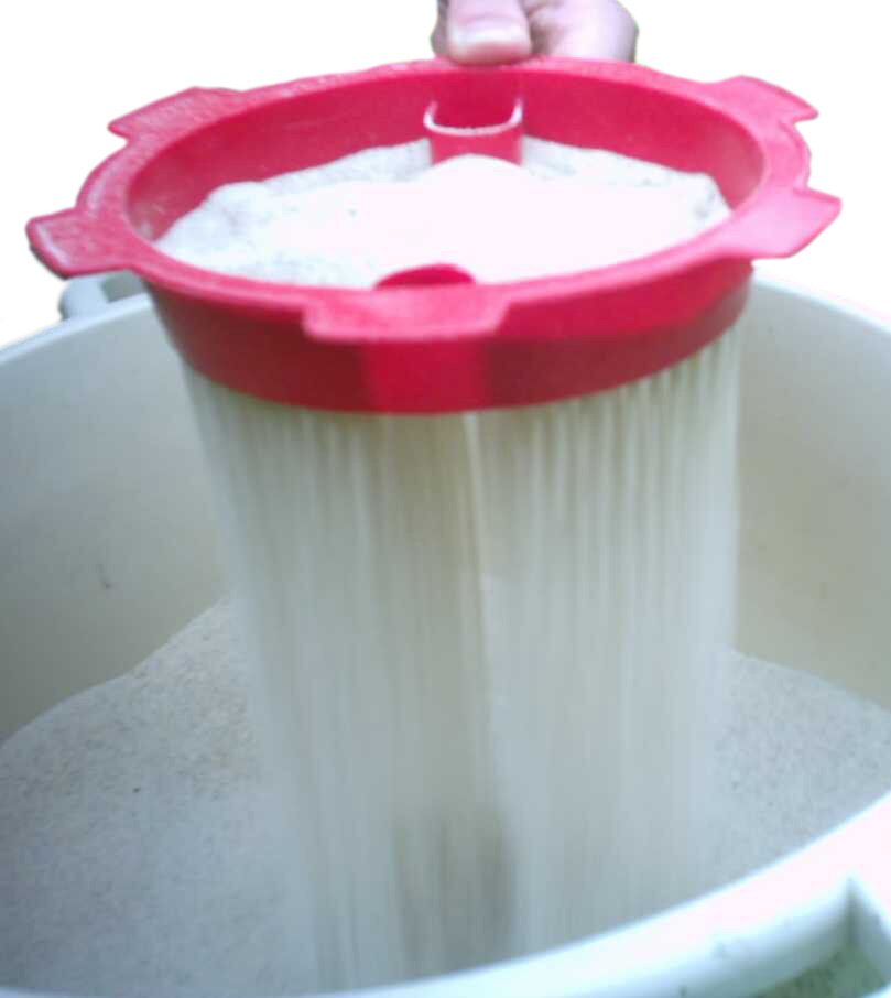 Zdjęcie przedstawia przesiewanie mąki. Widoczny jest przesiewacz ręczny – okrągłe czerwone sitko z prostokątnymi wypustkami na zewnętrznej krawędzi. W sitku znajduje się biała mąka, która przelatuje poniżej do białej miski, częściowo wypełnionej mąką.