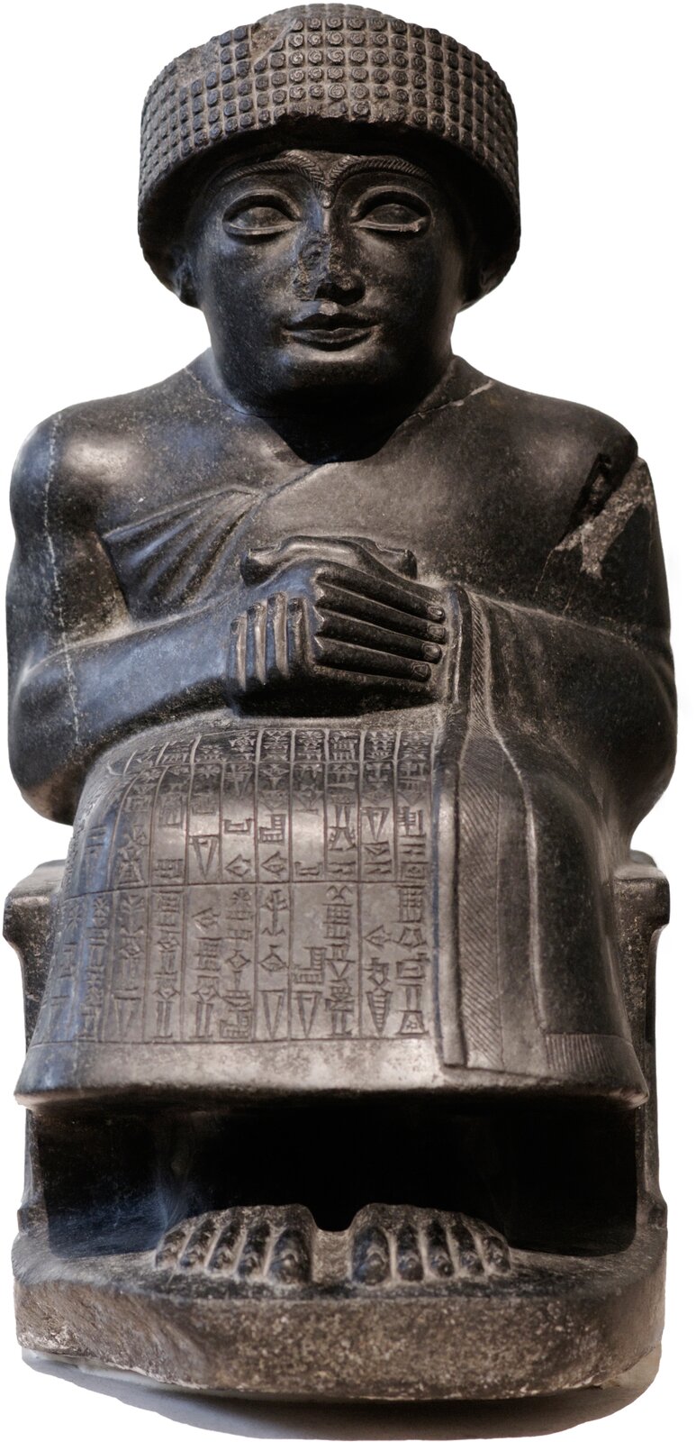Zdjęcie posągu przedstawiającego siedzącego na tronie masywnego mężczyznę. Ręce ma ugięte w łokciach i wsparte na kolanach, a dłonie trzyma skrzyżowane na piersi. Ubrany jest w nakrycie głowy oraz długą tunikę zasłaniającą nogi. Spod niej widoczne są wystające palce. Na przedniej stronie szaty i na tylnej części tronu widoczne są inskrypcje. 