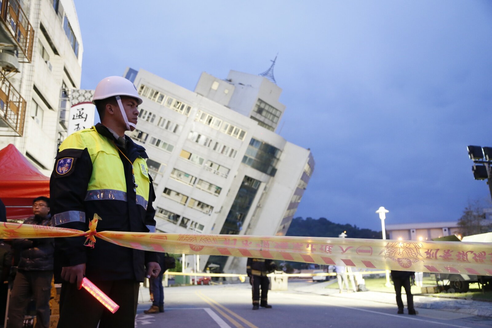 Zdjęcie ukazuje trzęsienie ziemi na Tajwanie. Po lewej stronie zdjęcia pracownik służb ratowniczych stoi za taśmą ostrzegawczą. W tle pochylony w prawo duży, kilkupiętrowy budynek.     