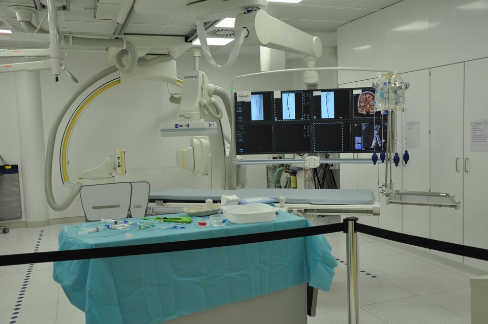 Zdjęcie przedstawia sale zabiegową w której znajduje się bardzo duże urządzenie. Nad stołem operacyjnym znajduje się osiem wielkich monitorów. 