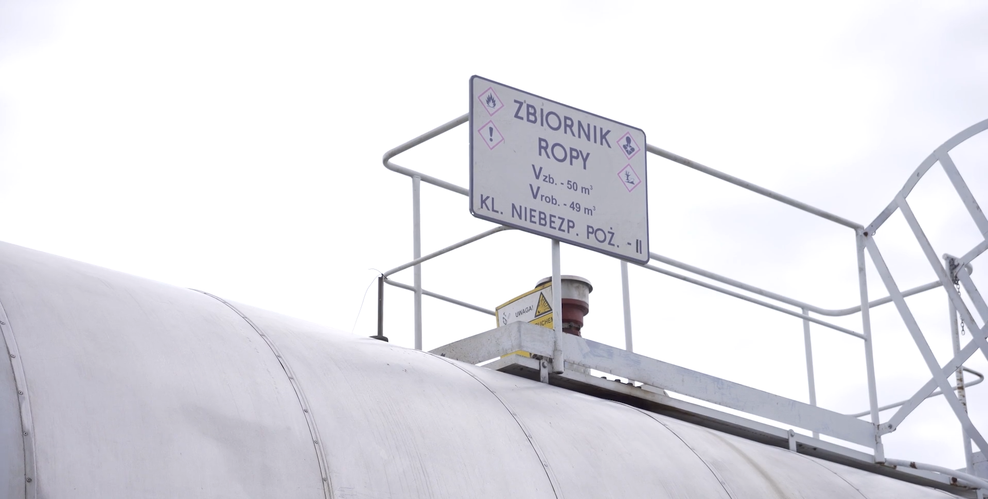 Zdjęcie przedstawia tablicę informacyjną wraz z górną częścią zbiornika magazynowego. Jest ona przymocowana do barierki, otaczającej platformę w górnej części cylindrycznego zbiornika. Treść tablicy jest następująca. Zbiornik ropy. Objętość zbiornika pięćdziesiąt metrów sześciennych. Objętość robocza zbiornika czterdzieści dziewięć metrów sześciennych. Klasa niebezpieczeństwa pożarowego druga. Na tablicy znajdują się również cztery piktogramy. Wszystkie składają się z czerwonej ramki białego wnętrza i czarnego symbolu we wnętrzu ramki. Po lewej stronie od napisu zamieszczono dwa piktogramy. Pierwszy przedstawia czarny płomień w czerwonej, kwadratowej ramce. Oznacza substancję łatwopalną. Drugi znajduje się poniżej i przedstawia znak wykrzyknienia. Co wskazuje na substancję o działaniu drażniącym. Po prawej stronie od napisu na tablicy zamieszczono dwa kolejne piktogramy. Pierwszy przedstawia zarys popiersia człowieka z rozchodzącym się promieniście dymem w klatce piersiowej. Piktogram oznacza działanie szkodliwe, rakotwórcze. Drugi, poniżej przedstawia brzeg rzeki, nad którym znajduje się drzewo pozbawione liści i martwa ryba. Oznacza on zagrożenie dla środowiska.