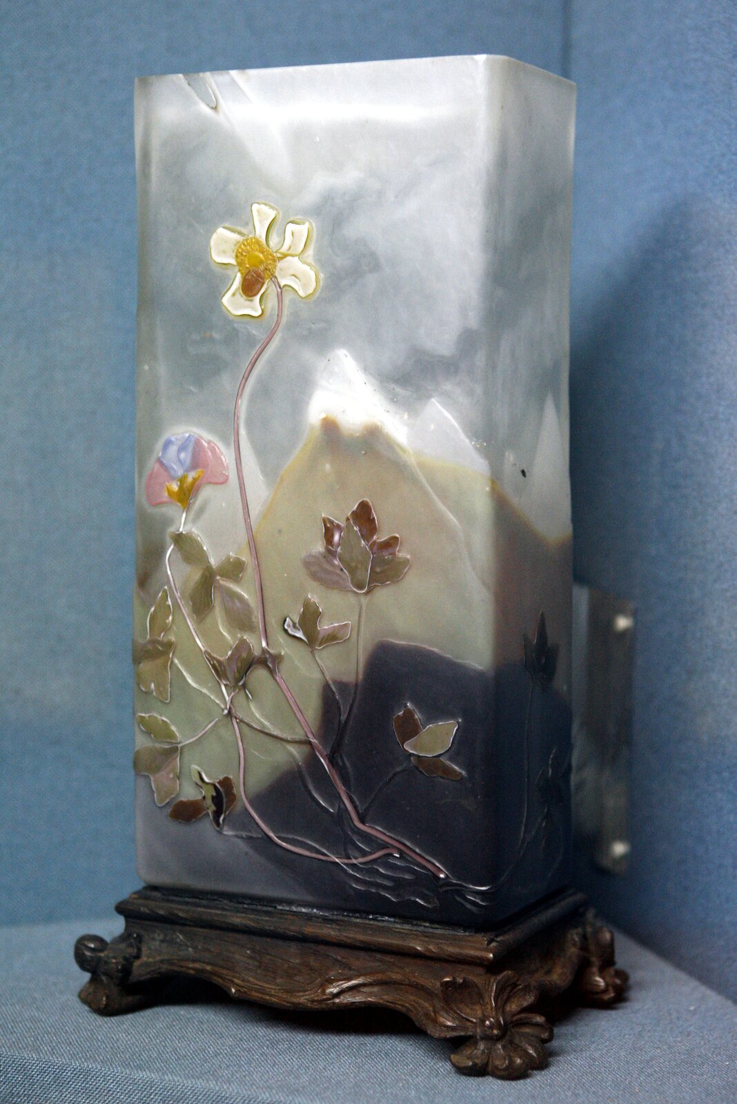 Ilustracja przedstawia wazon autorstwa Émile Gallé. Wazon ozdobiony jest motywami nawiązującymi do natury. Na pierwszym planie jest motyw roślinny, po czym w tle widać krajobraz górski. Wazon ma kształt prostokątny.