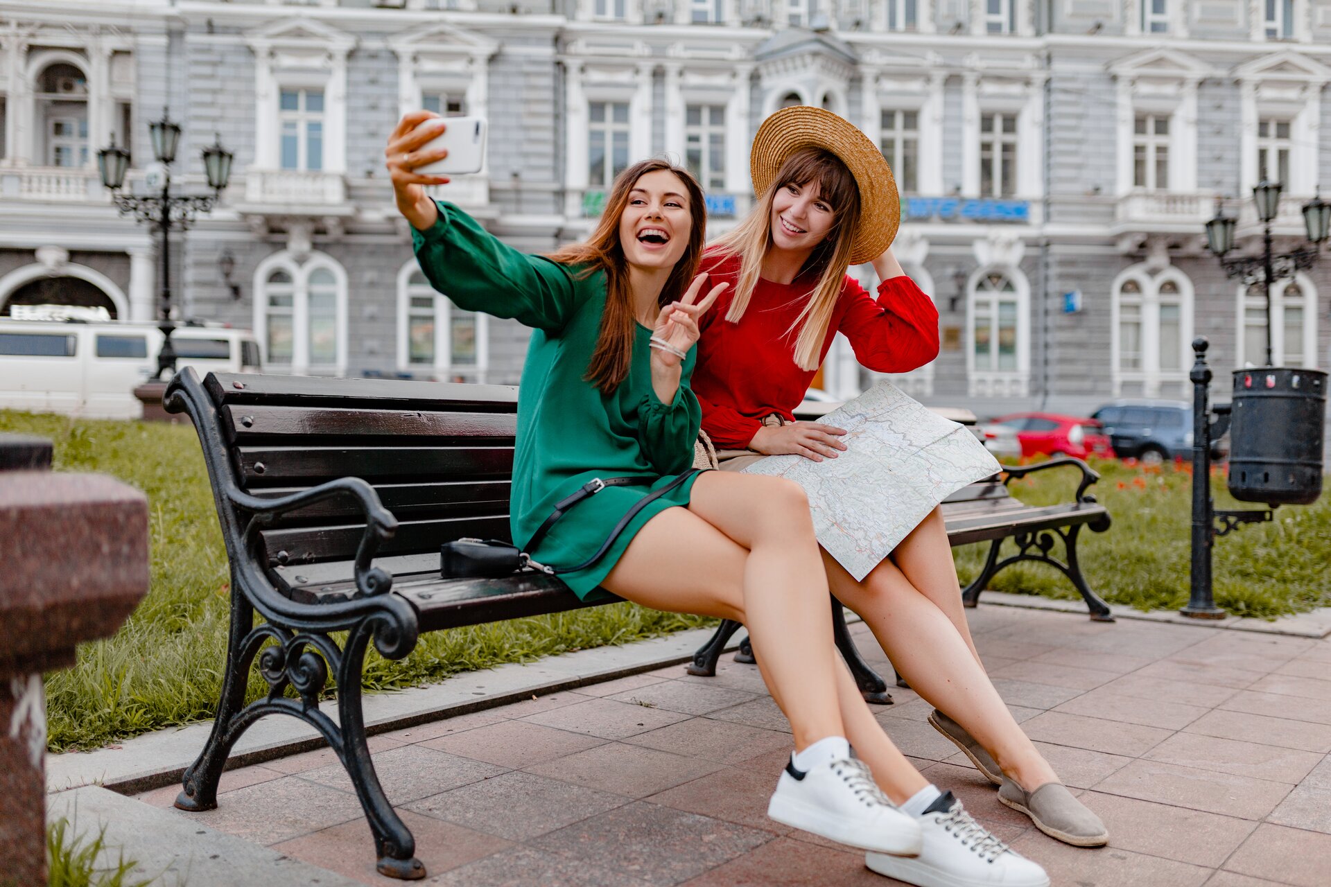 Zdjęcie przedstawia dwie młode uśmiechnięte dziewczyny, które siedzą na ławce i robią sobie selfie. 
W tle widoczny jest pas zieleni, ze stylowym koszem na śmieci oraz latarniami. Za pasem zieleni znajduje się elegancki budynek w stylu  secesyjnym.