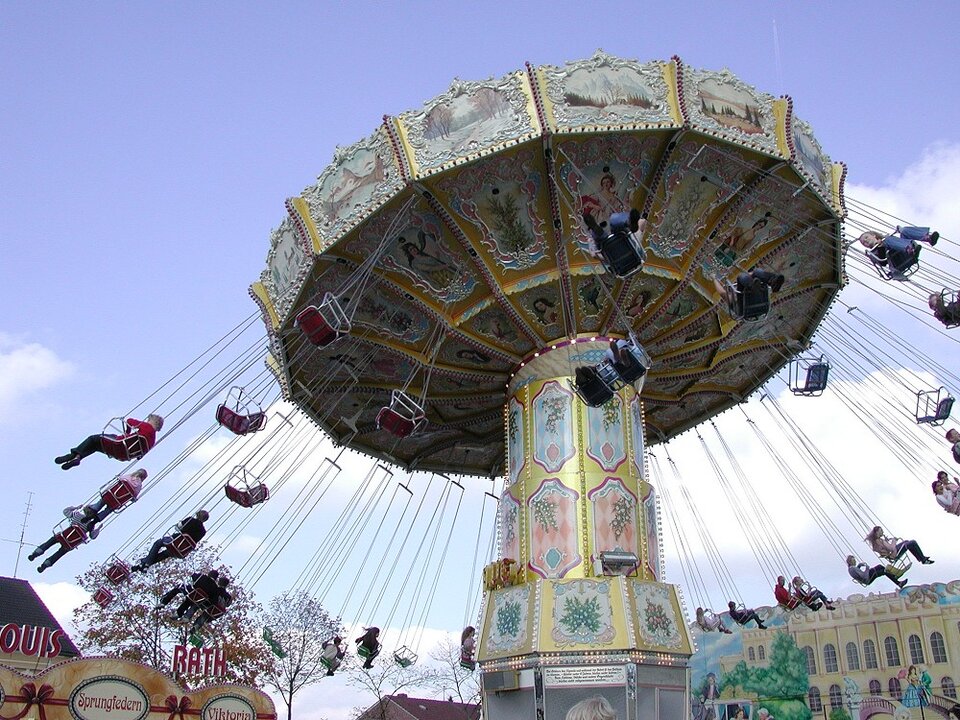 Zdjęcie przedstawia dużą karuzelę z siedziskami zawieszonymi na długich łańcuchach. Pasażerowie na siedziskach odchylają się na zewnątrz karuzeli. 
