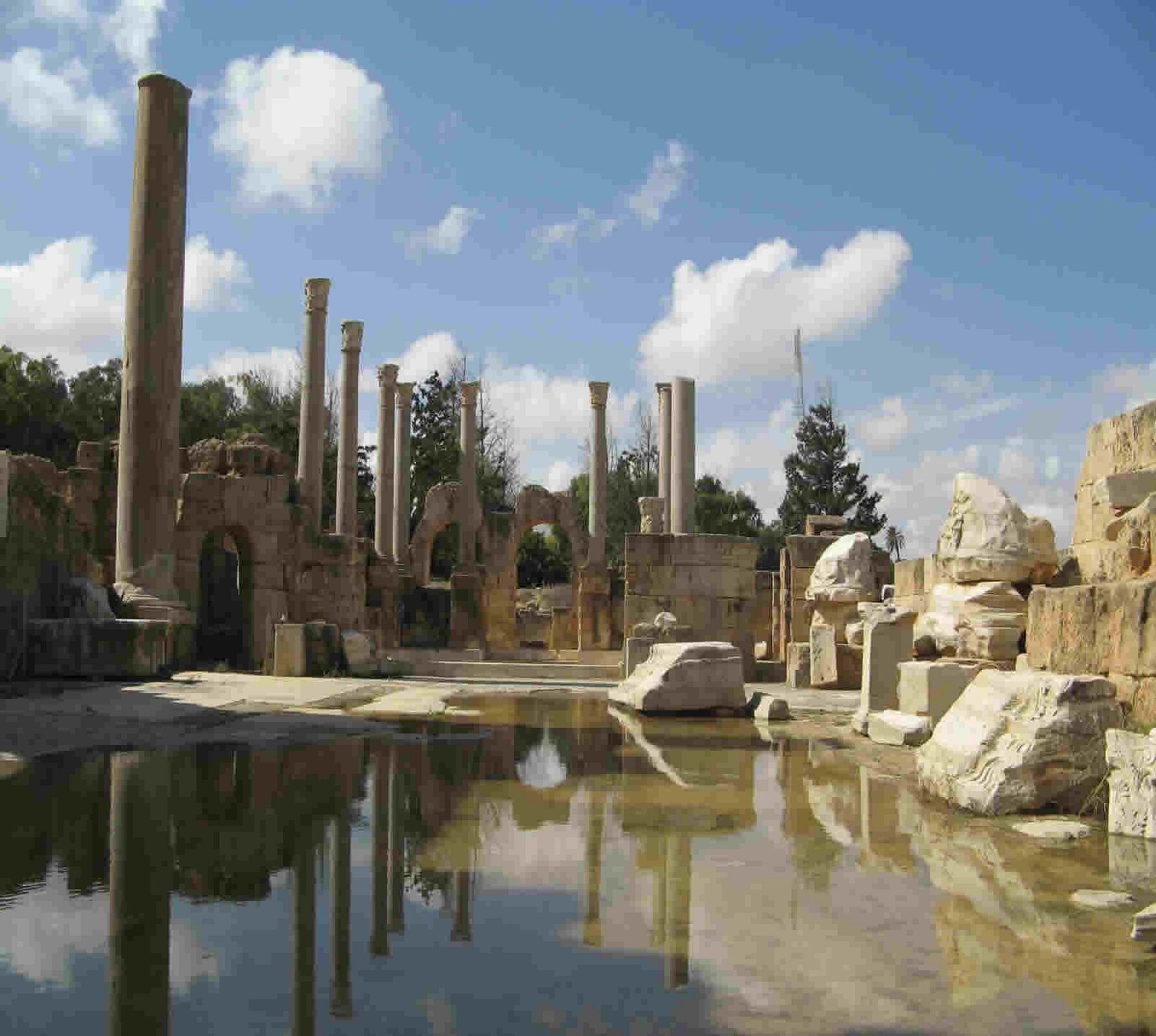 Fotografia nieznanego autora przedstawia widok na ruiny łaźni Hadriana, które znajdują się na dzisiejszym terytorium Libii. Na fotografii widoczne są kolumny oraz pozostałości murów budynku. Fotografia ruin została wykonana w pochmurny dzień, w tle widoczne są drzewa.