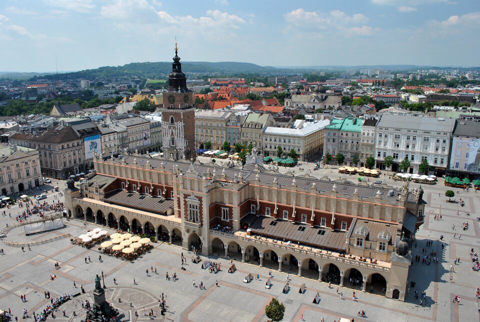 Ilustracja przedstawia widok z lotu ptaka na Krakowskie Sukiennice. Za rynkiem rozciąga się panorama miasta, na horyzoncie widoczne są wzgórza. Przed Sukiennicami przechadzają się ludzie.