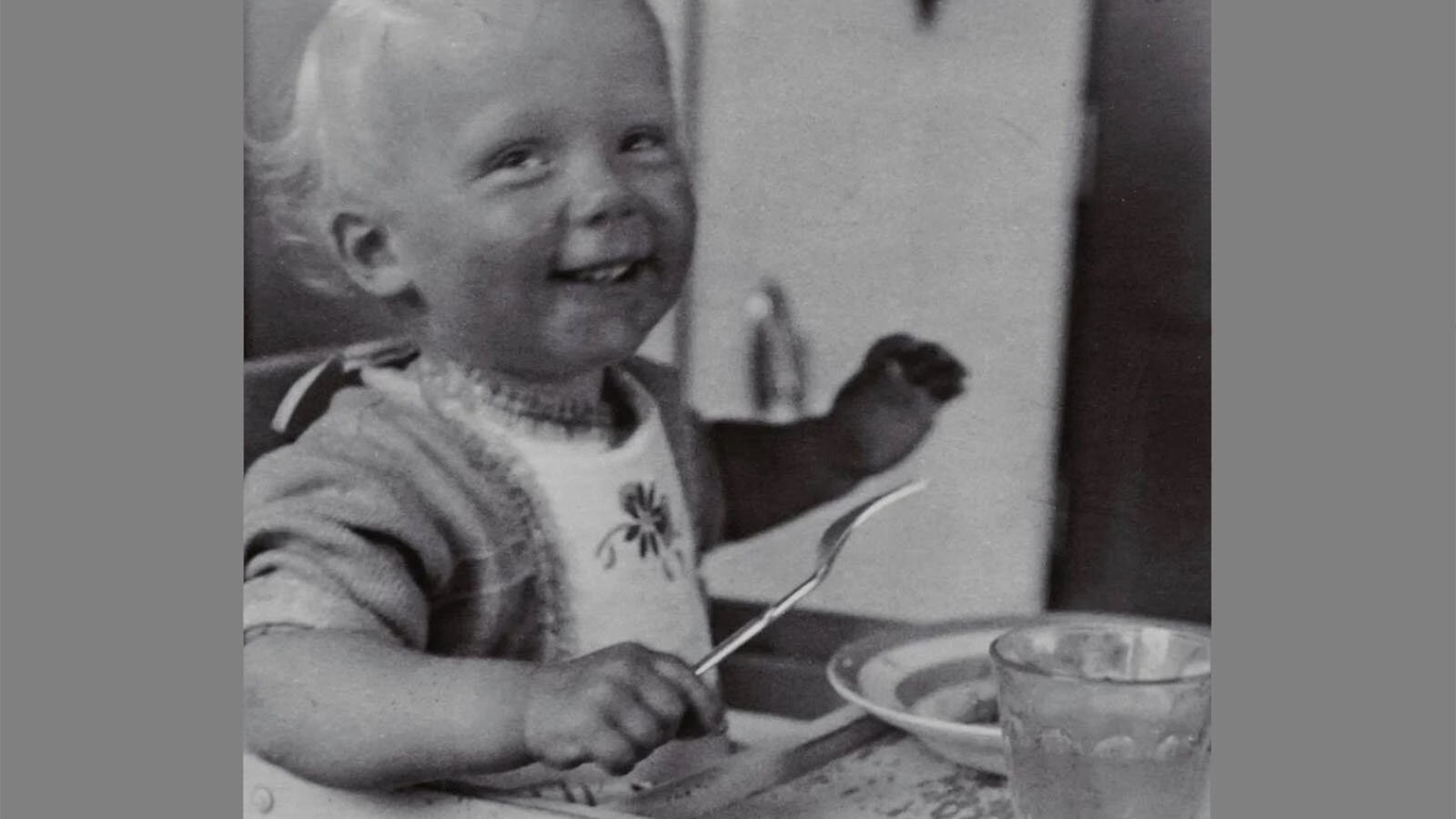 Czarno‑białe zdjęcie przedstawia uśmiechnięte małe dziecko, które siedzi przy stole. Dziecko ma buzię zwróconą w kierunku obiektywu, w prawej dłoni trzyma widelec, a lewą ma podniesioną do góry. Na piersi ma zawiązany śliniaczek. Na stole widoczny jest talerz oraz szklanka.