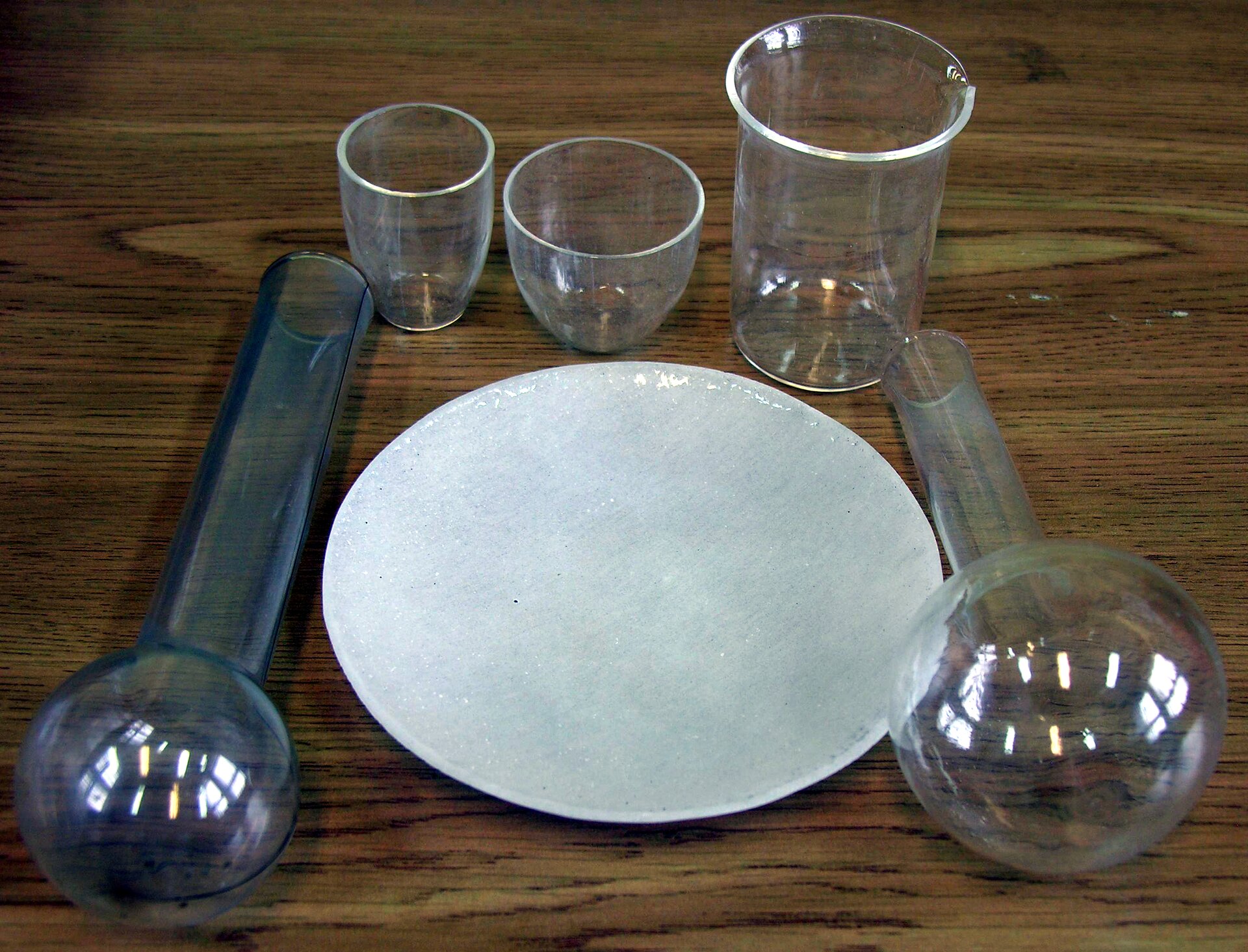 Zdjęcie przedstawia szklane naczynia laboratoryjne. Dwie leżące płasko kolbki kuliste z okrągłymi szyjkami, pomiędzy nimi talerzyk z błyszczącym szkliwem, powyżej trzy naczynia stojące, w tym dwa tygielki i jedna zlewka.