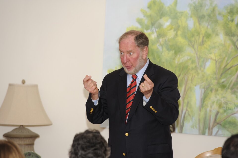 Ilustracja przedstawia mężczyznę w średnim wieku w czasie wykładu. Mężczyzna stoi koło katedry, przed nim znajdują się rzędy krzeseł dla studentów. W drugim rzędzie siedzi studentka zwrócona twarzą do przodu do wykładowcy.