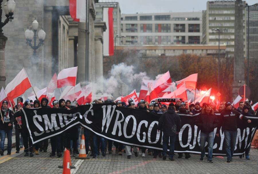 Zdjęcie przedstawia tłum młodych mężczyzn maszerujących w pochodzie w pobliżu Pałacu Kultury w Warszawie. Mężczyźni w pierwszym rzędzie pochodu niosą czarny baner z białym napisem Śmierć wrogom ojczyzny. Uczestnicy pochodu niosą liczne polskie flagi oraz trzymają odpalone race. Nad nimi unosi się dym. Na budynku Pałacu Kultury zawieszone są polskie flagi.
