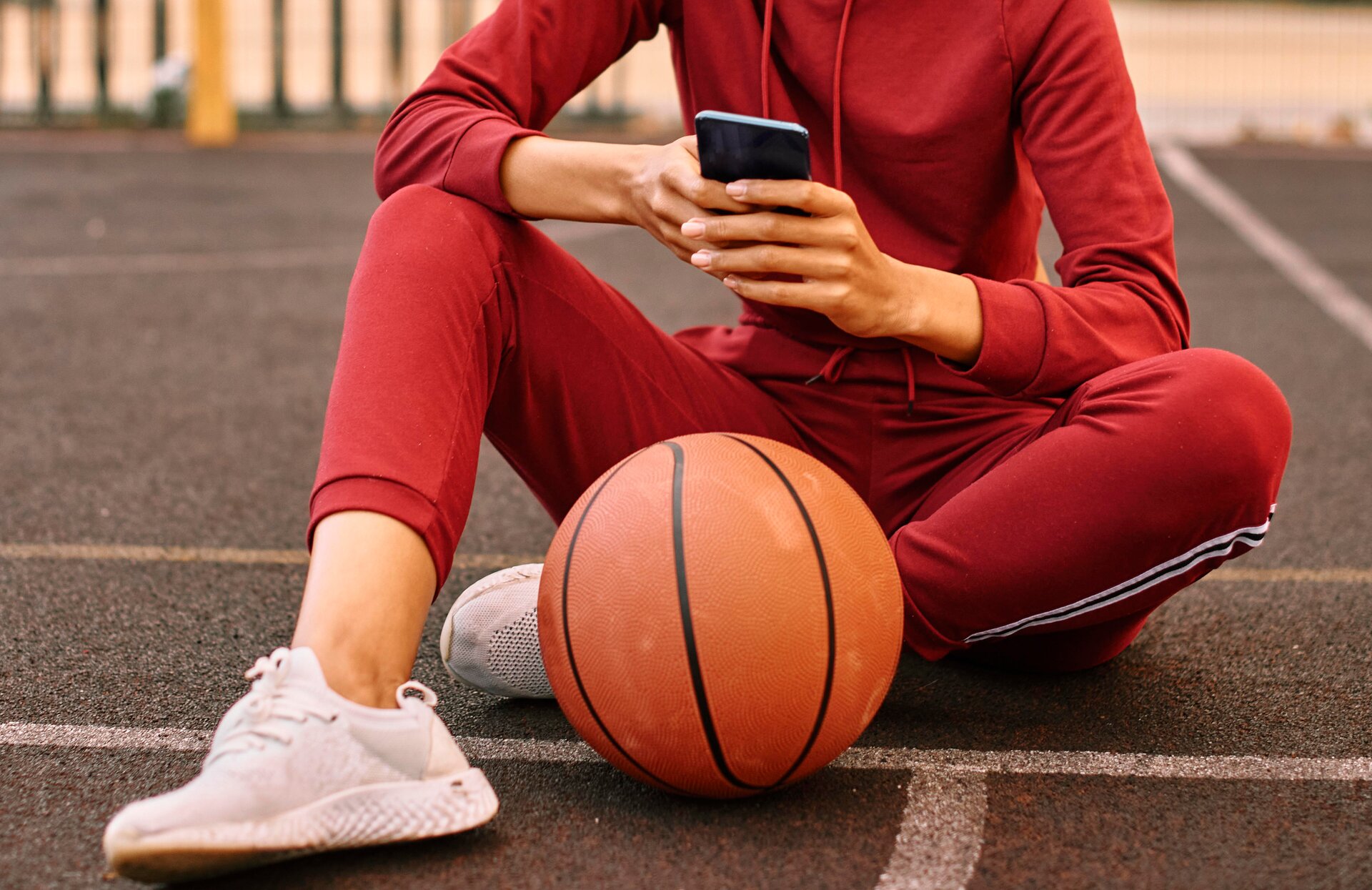 Zdjęcie przedstawia postać, w ujęciu od ramion w dół, która siedzi na boisku. Postać jest ubrana w czerwony dres oraz białe sportowe obuwie. Prawa noga jest zgięta w kolanie, na którym oparta jest prawa ręka. Lewa noga jest zgięta w kolanie w takim sposób, że dotyka podłoża, a lewa stopa jest pociągnięta pod prawe udo. Przed stopami widoczna jest piłka do koszykówki. Postać trzyma telefon komórkowy w dłoniach.