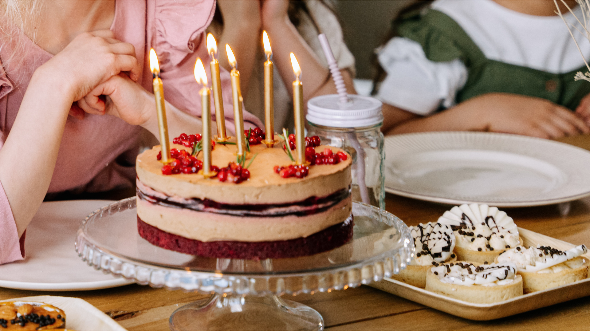 Zdjęcie przedstawia na pierwszym planie zbliżenie na tort urodzinowy z sześcioma płonącymi świeczkami. Tort znajduje się na paterze. Obok na talerzyku znajdują się słodkie babeczki. Na drugim planie widoczne są ręce osób, które siedzą przy stole. Przed nimi stoją puste talerzyki.
