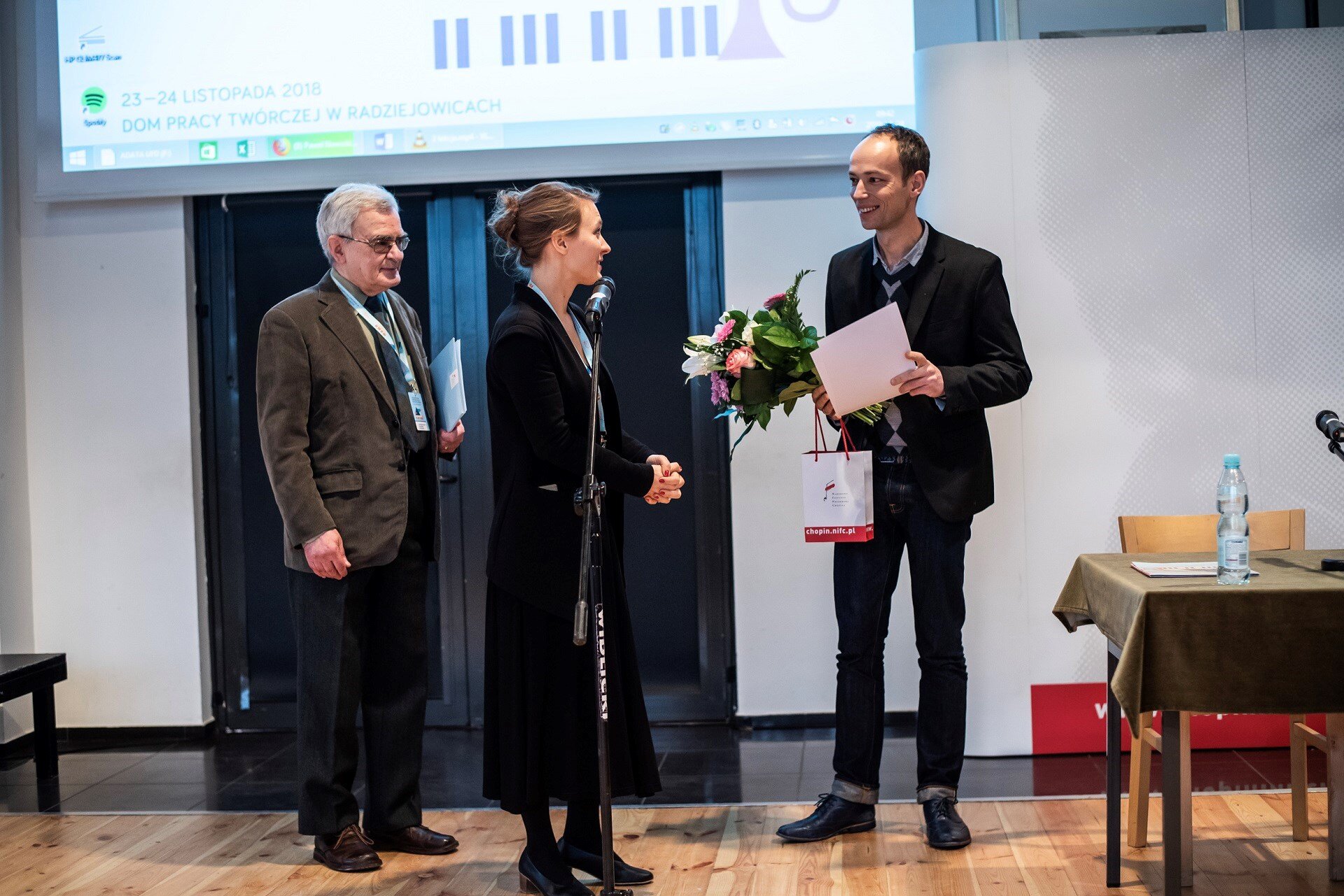 Fotografia przedstawia troję ludzi stojących na scenie. Dwóch mężczyzn oraz kobieta stojąca pośrodku. Jeden z mężczyzn jest w podeszłym wieku, ma siwe włosy oraz okulary na nosie. Ubrany jest w brązową marynarkę oraz ciemne garniturowe spodnie. Stojąca obok niego młoda ciemnowłosa kobieta ubrana jest w długi czarny płaszcz. Spogląda się na młodego łysiejącego mężczyznę ubranego w garnitur. Mężczyzna ten trzyma w dłoniach kwiaty, arkusz papieru oraz torebkę. 