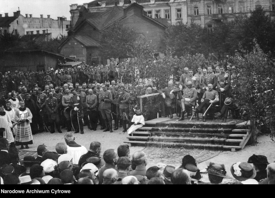 Zdjęcie przedstawia ludzi zgromadzonych na dużym placu w mieście. Pośrodku placu stoi trybuna, zasiadają na niej wojskowi notable: Józef Piłsudski i generałowie, a za ich plecami stoją wojskowi niżsi stopniem. Wokół trybuny zgromadzony jest tłum cywilnych ludzi, księża oraz duża grupa wojskowych. W tle zabudowania mieszkalne, wysokie kamienice. 
