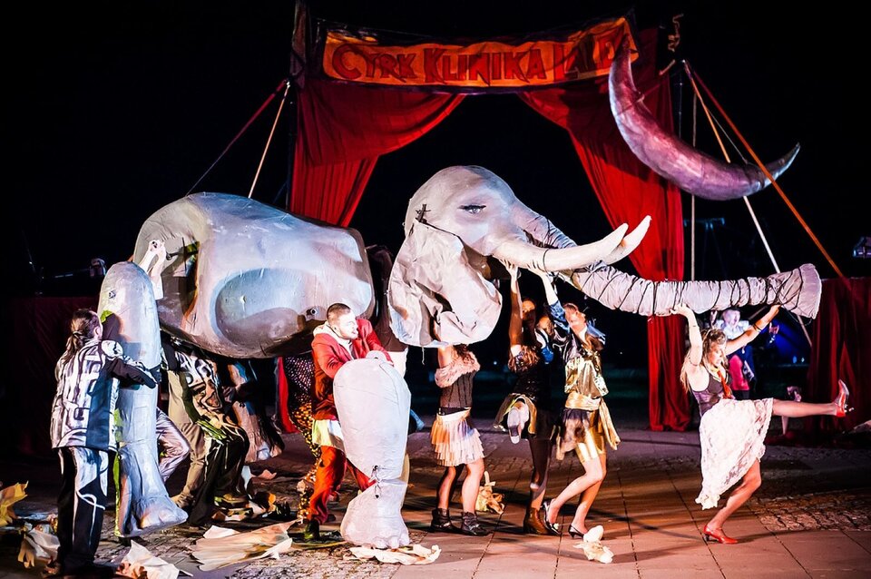 Zdjęcie przedstawia scenę ze spektaklu plenerowego Teatru "Klinika Lalek",  który odbył się podczas XXII Międzynarodowego Festiwalu Sztuka Ulicy w Warszawie. To dynamiczny pochód bajecznie ubranych aktorów idących ze słoniem. Słoń jest wielką lalką i składa się z kilku ogromnych części animowanych przez ludzi. Prowadzi kobieta krokiem defiladowym, podtrzymując długą trąbę słonia. Za nimi stoi konstrukcja z kurtyną, na której został zawieszony połyskujący księżyc, na niej transparent z napisem: "Cyrk Klinika Lalek". Spektakl odbywa się nocą na oświetlonym placu miejskim, nosi tytuł: "Cyrk bez przemocy". Wszyscy bawią się doskonale.