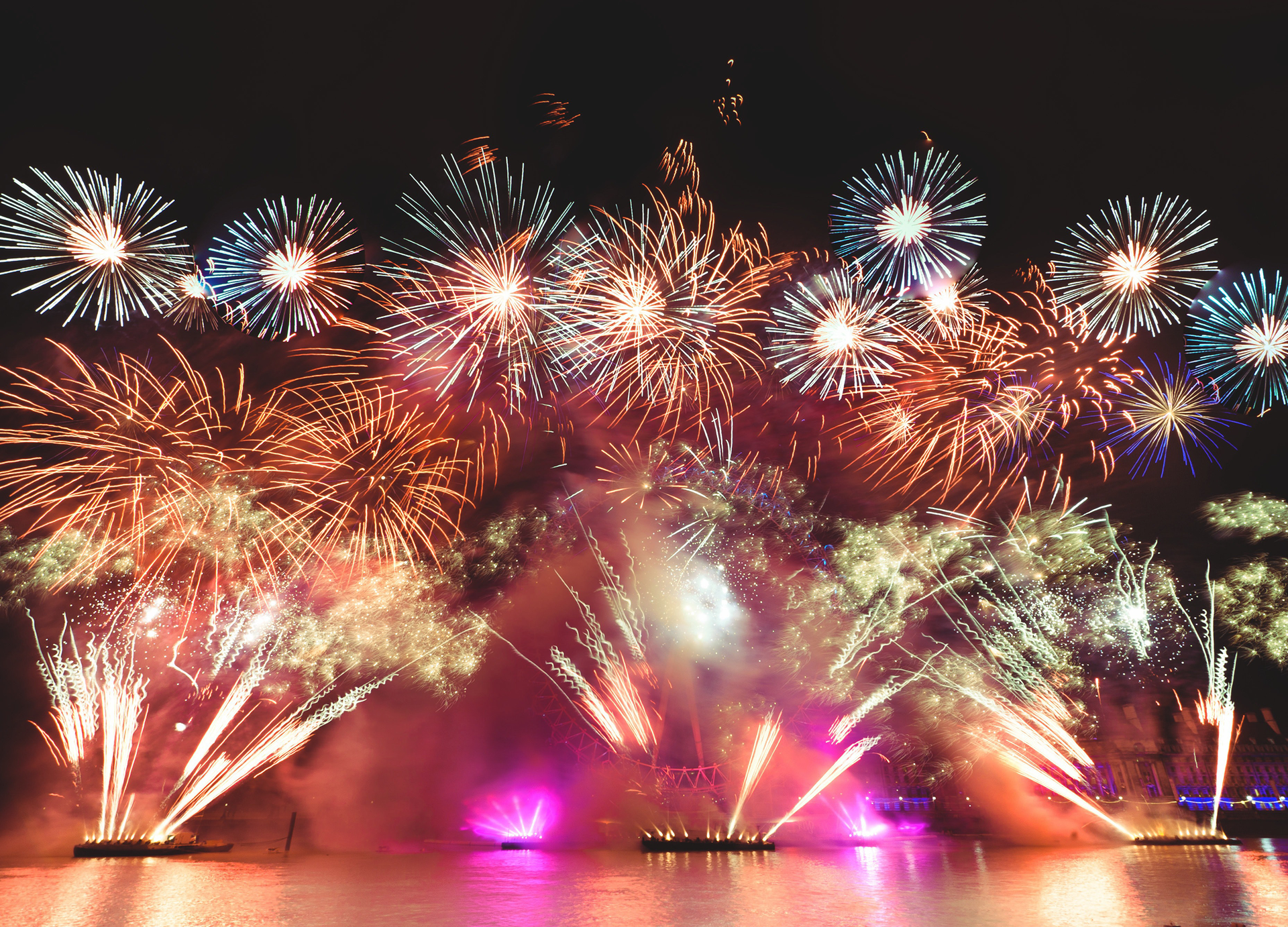Zdjęcie przedstawia nocny pokaz fajerwerków nad brzegiem zbiornika wodnego. Na pierwszym planie widoczna jest tafla wody, w której odbijają się sztuczne ognie. Na dalszym planie widoczne są kolorowe fajerwerki na dwóch poziomach, tuż nad taflą wody i na ciemnym niebie.