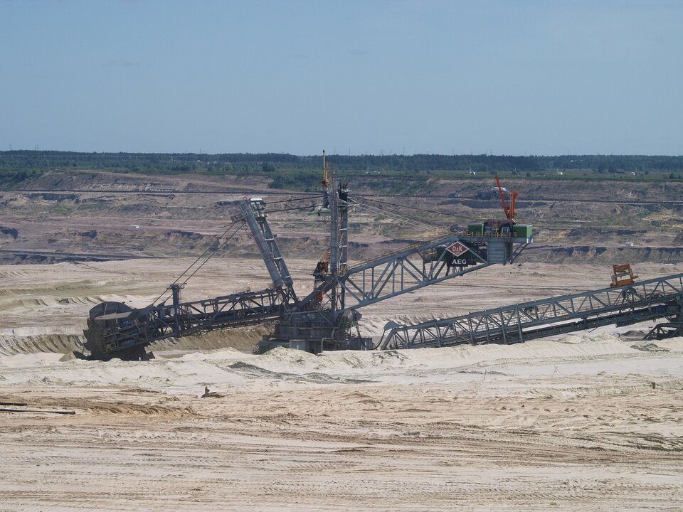 Zdjęcie przedstawia teren, na którym pracuje ciężka maszyna. Maszyna jest w dołku. Teren wokół jest eksploatowany, ma wzniesienia i obniżenia w postaci schodów, tarasów.  
