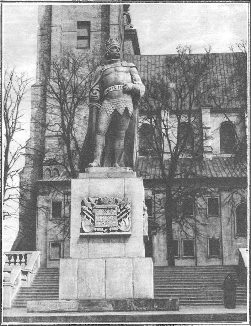 Zdjęcie przedstawia pomnik króla stojący na wysokim cokole. Mężczyzna ma na głowie koronę, na ramionach płaszcz, w pasie pas, w prawym ręku miecz.