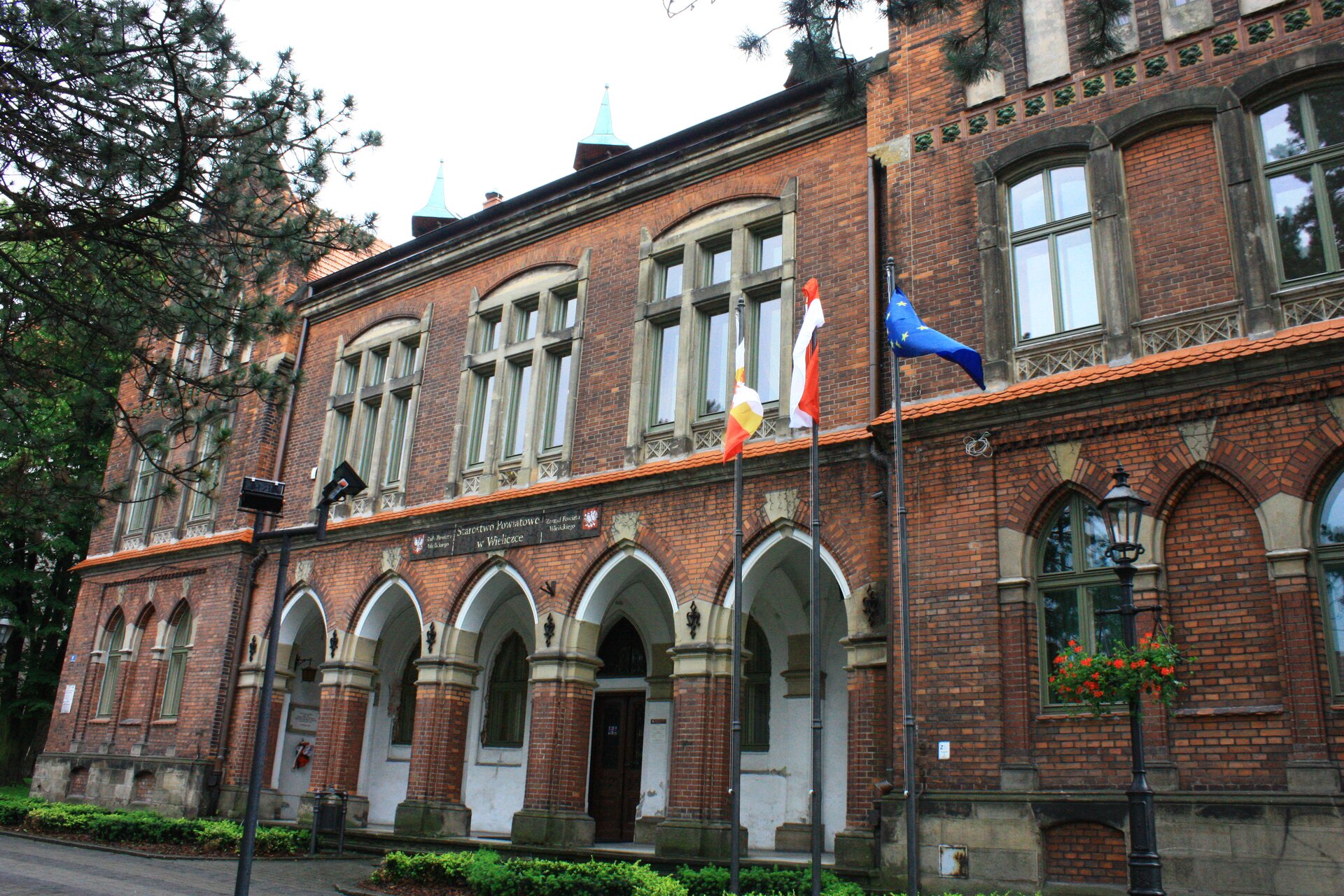 Kliknij żeby powiększyć Na zdjęciu znajduje się zabytkowy budynek - siedziba starostwa wielickiego w Wieliczce. Przed wejściem do budynku są charakterystyczne strzeliste łuki. Okna umieszczone po bokach są również zakończone łukami.