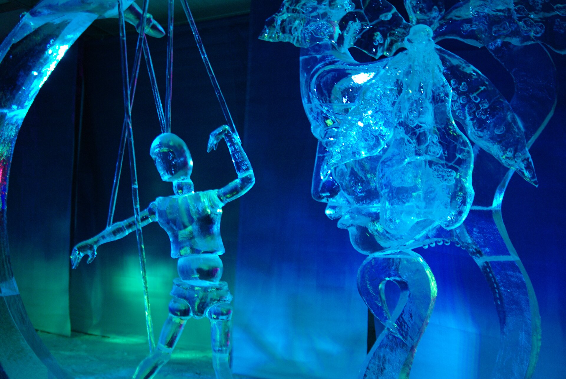 Ilustracja przedstawia rzeźby wykonane z lodu. Z lewej strony znajduje się rzeźba kukiełki na sznurkach, a po prawej stronie rzeźba twarzy. W pomieszczeniu, w którym prezentowane są rzeźby oświetlenie jest niebieskie i zielone. Taki sam kolor mają rzeźby.