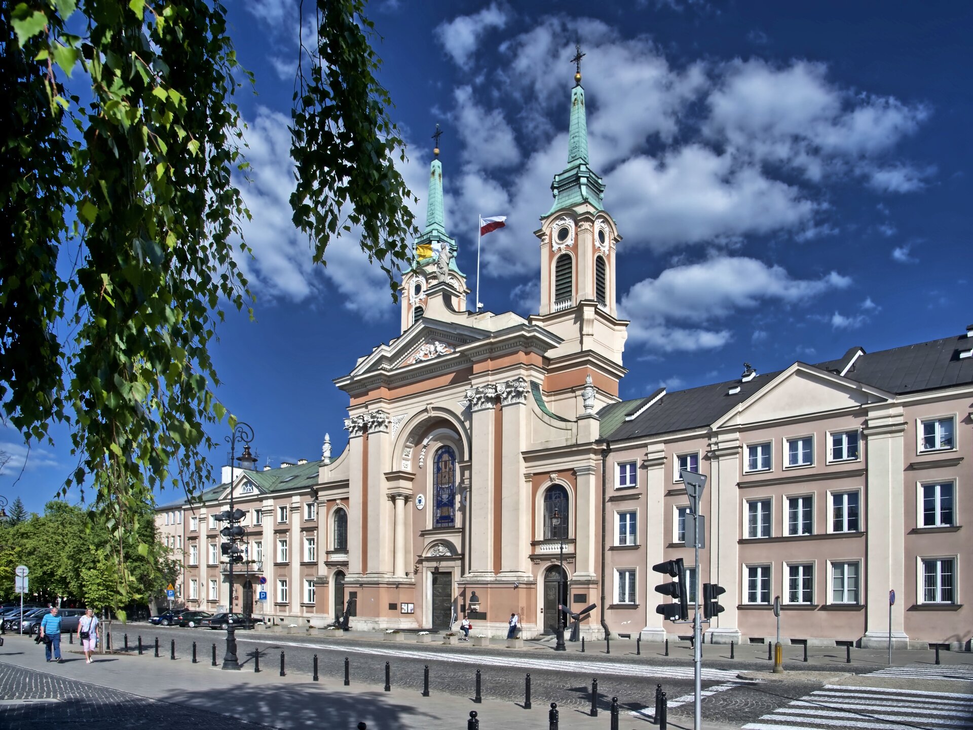 Fotografia przedstawiająca Kościół pijarów w Warszawie, dziś Katedra Polowa Wojska Polskiego, 1758–1769. Budynek ma jasną elewację i strzeliste dwie wieże zakończone krzyżami. Na dachu znajduje się maszt z flagą Polski.