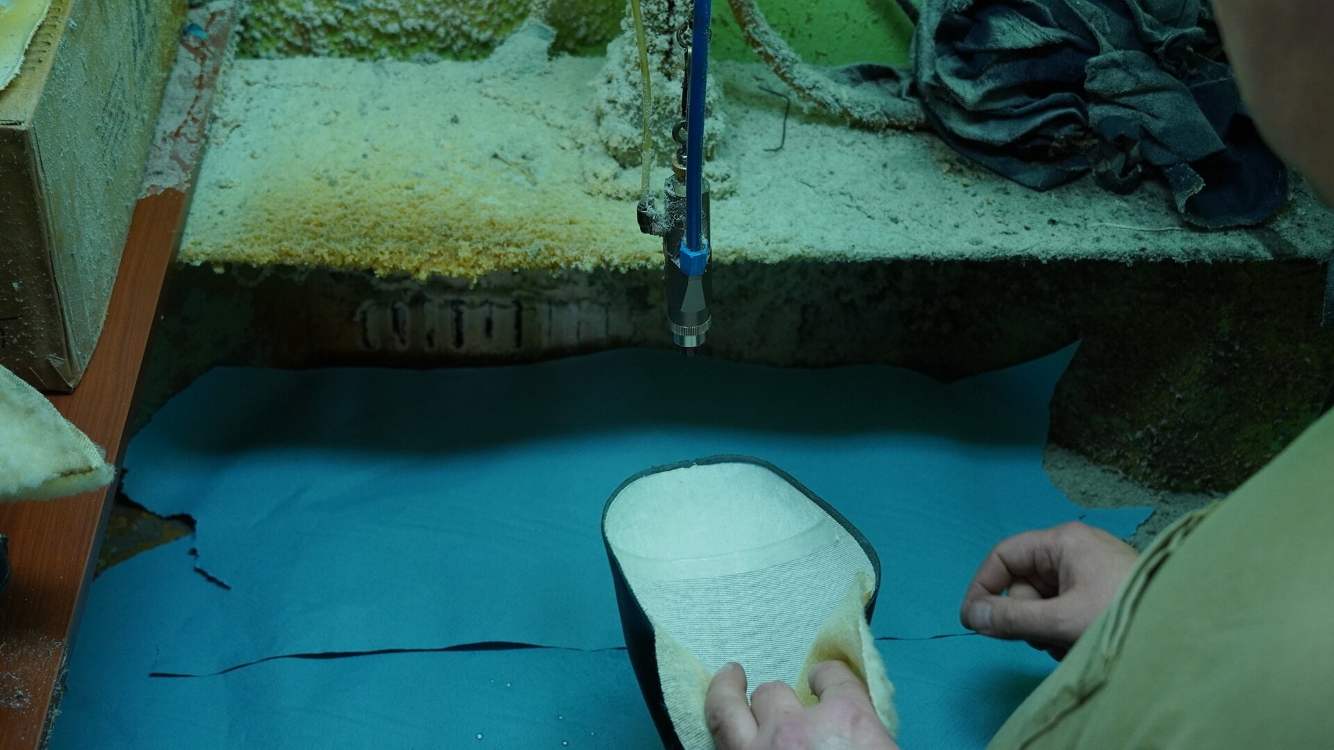 Zdjęcie przedstawia widok z góry na stanowisko pracy, tkaninę, na której znajduje się element cholewki przytrzymywany przez pracownika i podkładany pod rurkę, która stanowi część urządzenia służącego do nakładania kleju. Nagranie tożsame z treścią.