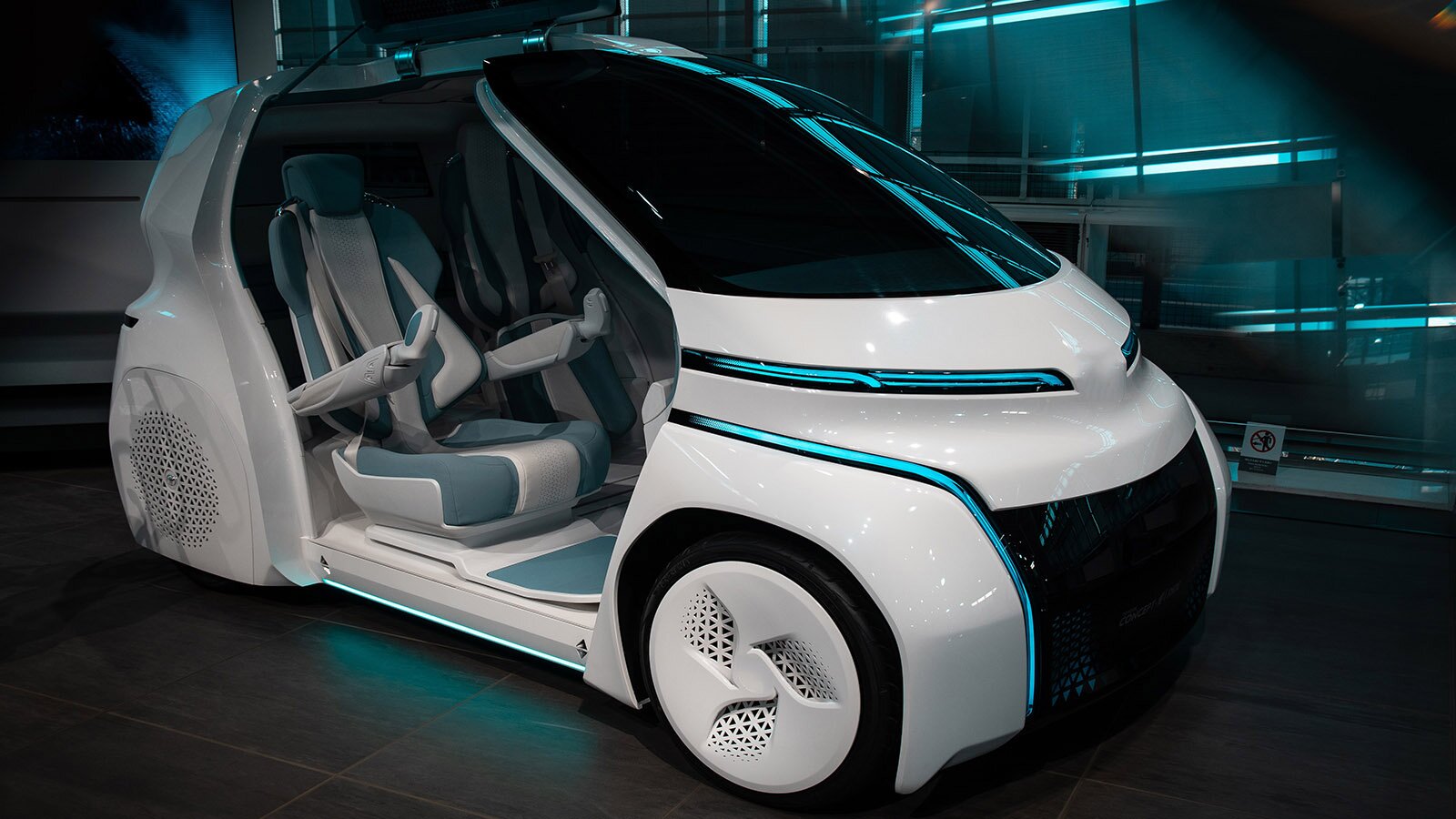 Zdjęcie przedstawia futurystyczny, samojeżdżący samochód. Samochód jest zaprezentowany do sprzedaży w salonie samochodowym. Auto ma otwarte drzwi ukazując nowoczesne wnętrze samochodu.  