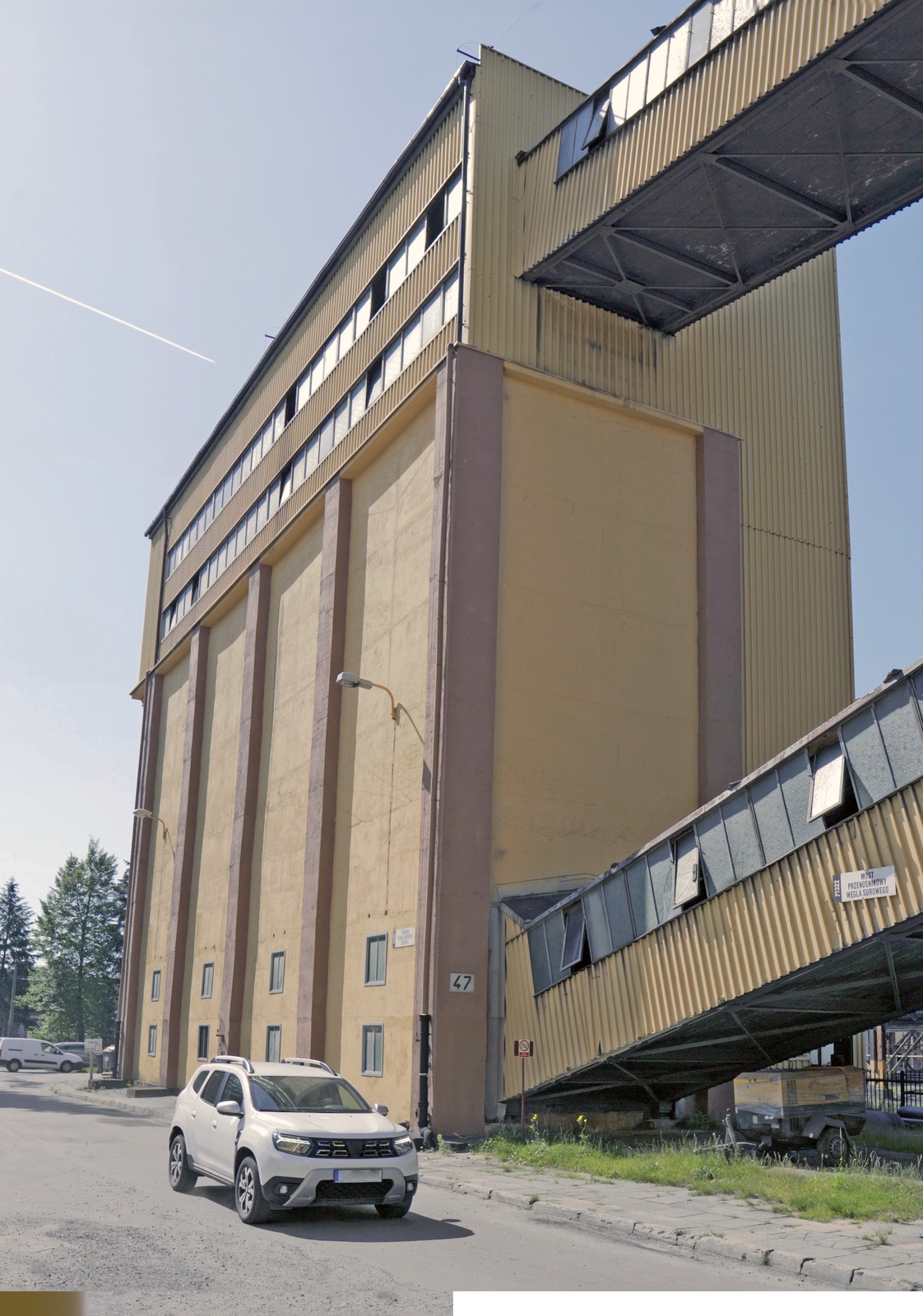 Na zdjęciu widoczny jest teren przemysłowy pokazany dołu. Po lewej stronie zdjęcia widoczny jest duży prostopadłościenny budynek. Do podstawy budynku przyłączony jest łącznik. Łącznik znajduje się pod kątem. Łącznik wykonany jest z żółtej blachy falistej. W górnej części ściany osadzone są okna. Na boku łącznika widoczna jest tablica z napisem: most przenośnikowy węgla surowego. Przy budynku zaparkowany jest samochód.Na terenie placu widać części urządzeń oraz metalową konstrukcję przypominającą ramę przenośnika.