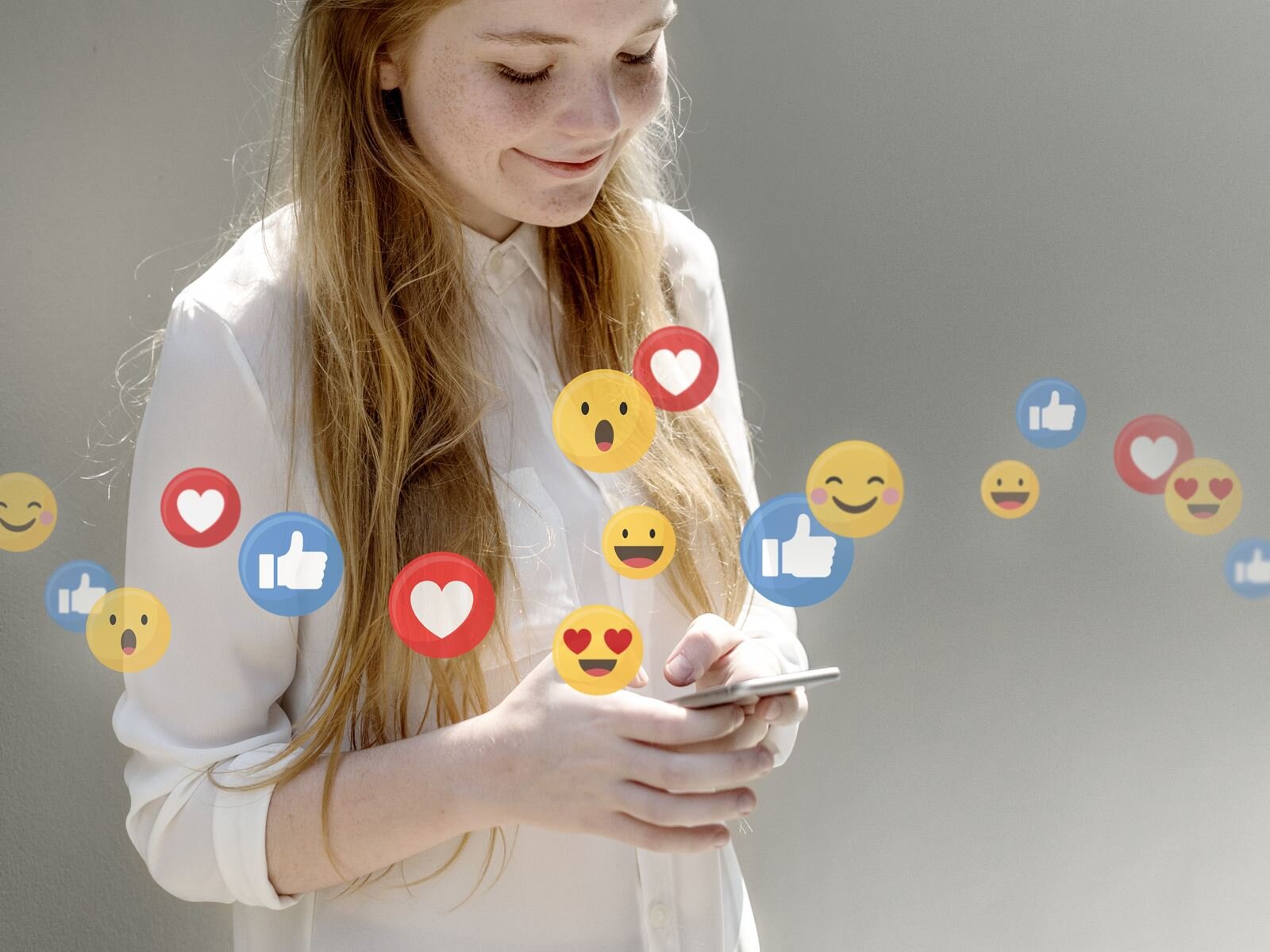 Zdjęcie przedstawia nastoletnią, uśmiechającą się dziewczynę od pasa w górę, zwróconą w prawo. Jej głowa jest lekko pochylona, a wzrok skierowany na ekran telefonu, który trzyma w dłoniach. Dziewczyna ma długie, rozpuszczone blond włosy i jest ubrana w białą koszulę. Na wysokości klatki piersiowej dziewczyny, przez całą szerokość zdjęcia, widoczne są emotikonki, w tym: serce, kciuk w górę(like) oraz buźki symbolizujących zdziwienie, śmiech, uśmiech, polubienie.