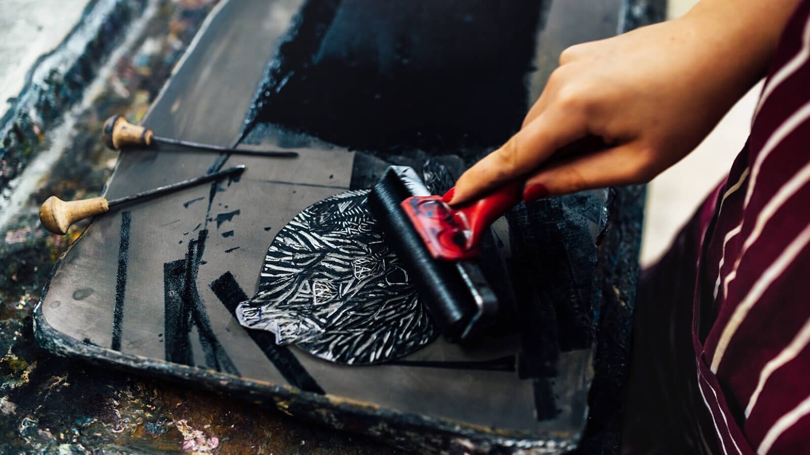 Fotografia przedstawia widok na nakładanie farby drukarskiej na linoleum. Na stole znajduje się plastikowa wanna, w której leżą dwa rylce oraz wzornik kształtem przypominający liść, na który nakładana jest czarna farba za pomocą wałka o czerwonym trzonie. Wałek trzymany jest przez kobietę, która, oprócz dłoni z pomalowanymi na czerwono paznokciami oraz fragmentu czerwonego fartucha w białe paski, jest niewidoczna na fotografii. 