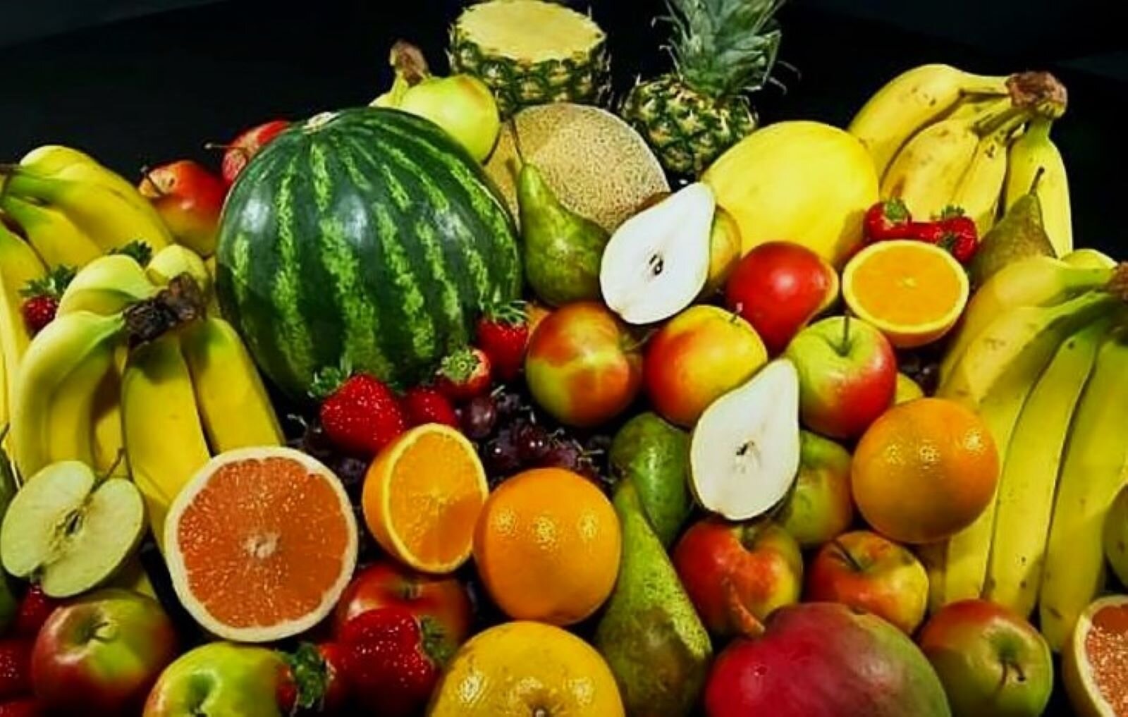 Fotografia przedstawia stertę owoców w dużym zbliżeniu. Bez trudu można rozpoznać jabłka, pomarańcze, truskawki, gruszki, banany. Wyróżnia się wielki arbuz i dwa ananasy, niektóre owoce są przekrojone. Jest to fotografia reklamowa.