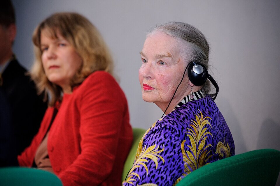 Na fotografii widoczna jest Jadwiga Staniszkis siedząca przy stole podczas konferencji naukowej. Ujęcie pokazuje ją od pasa w górę. Ma upięte z tyłu siwe włosy, ubrana jest we wzorzystą, elegancką koszulę (złote motywy liści na fioletowym tle). Patrzy przed siebie, jakby słuchała wypowiedzi. Uwagę zwraca jej czerwona szminka na ustach. W tle znajduje się druga kobieta, częściowo widoczna i nieostra, co sugeruje przestrzeń konferencyjną.