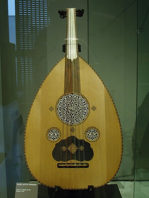 Fotografia przedstawia ustawioną pionowo lutnię arabską – strunowy instrument muzyczny. Jej płyta rezonansowa wykonana jest z jasnego drewna, a strunnik i podstrunnica z ciemnego. Posiada ona owalny kształt zwężający się w stronę gryfu, zakończonego główką z kluczami do naciągania strun. Jest ich dwanaście ułożonych po dwie od najcieńszych do najgrubszych. W każdej parze struny mają taką samą grubość. W płycie wierzchniej widzimy trzy otwory: na środku przy podstrunnicy jeden większy, a dwa mniejsze przy strunniku. Wszystkie są zamknięte ozdobną rozetą. Lutnia znajduje się w szklanej gablocie.
