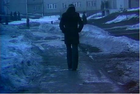 Zdjęcie przedstawia idącego samotnie mężczyznę. Ubrany jest w ciemną kurtkę i czapkę, ręce trzyma w kieszeni. Ma lekko pochyloną głowę. Na ziemi leżą pozostałości po śniegu. W tle stoi blokowisko. Po lewej znajduje się kilka postaci.